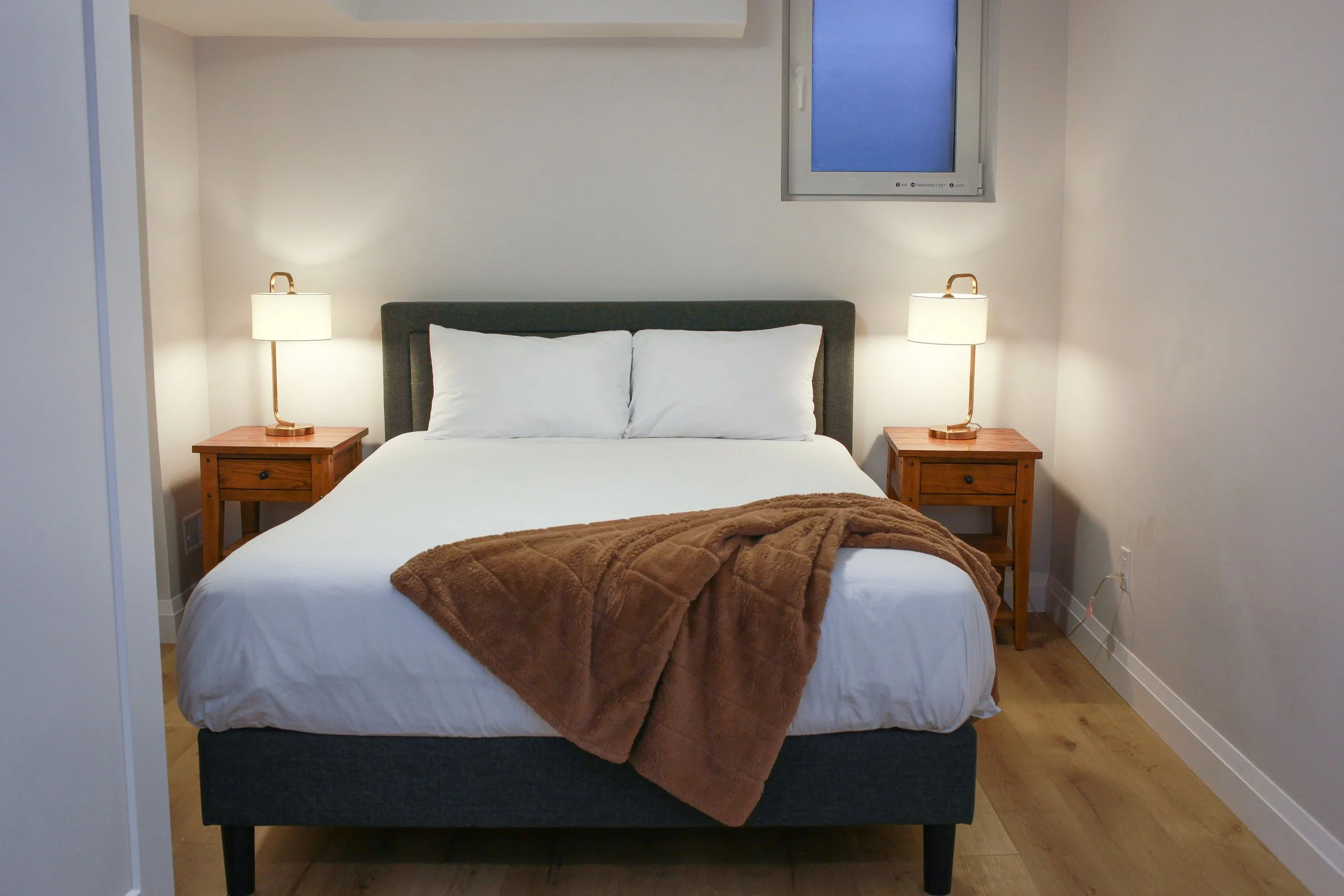 A neatly made bed with white sheets, two white pillows, and a brown blanket in a cozy bedroom. There are two wooden nightstands with lamps on either side of the bed, and a small window above the bed.
