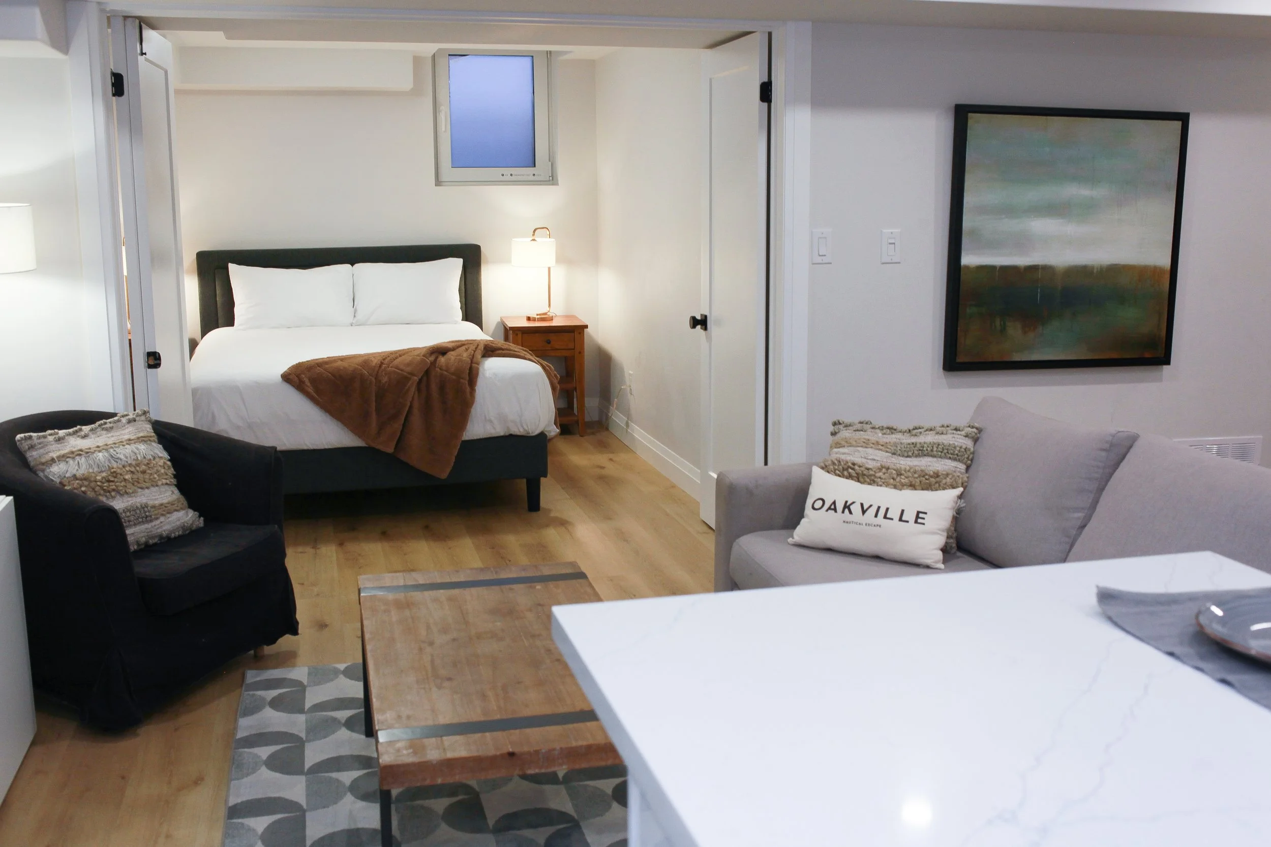Living space with a bedroom in the background and a sitting area in the foreground, including a sofa, armchair, wooden coffee table, artwork on the wall, and a bed with white linens and a brown throw blanket.