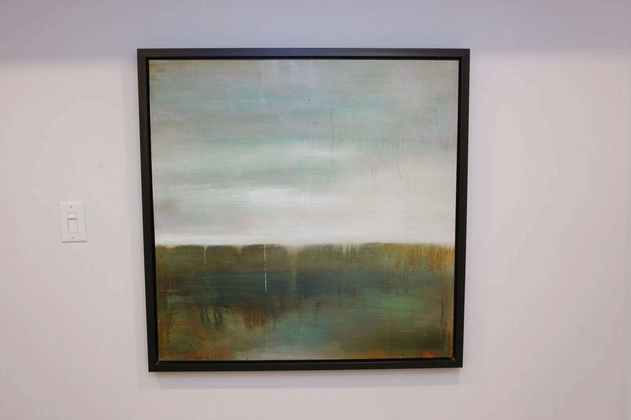 Abstract landscape painting with soft green, white, and brown tones, framed in black, hanging on a white wall.