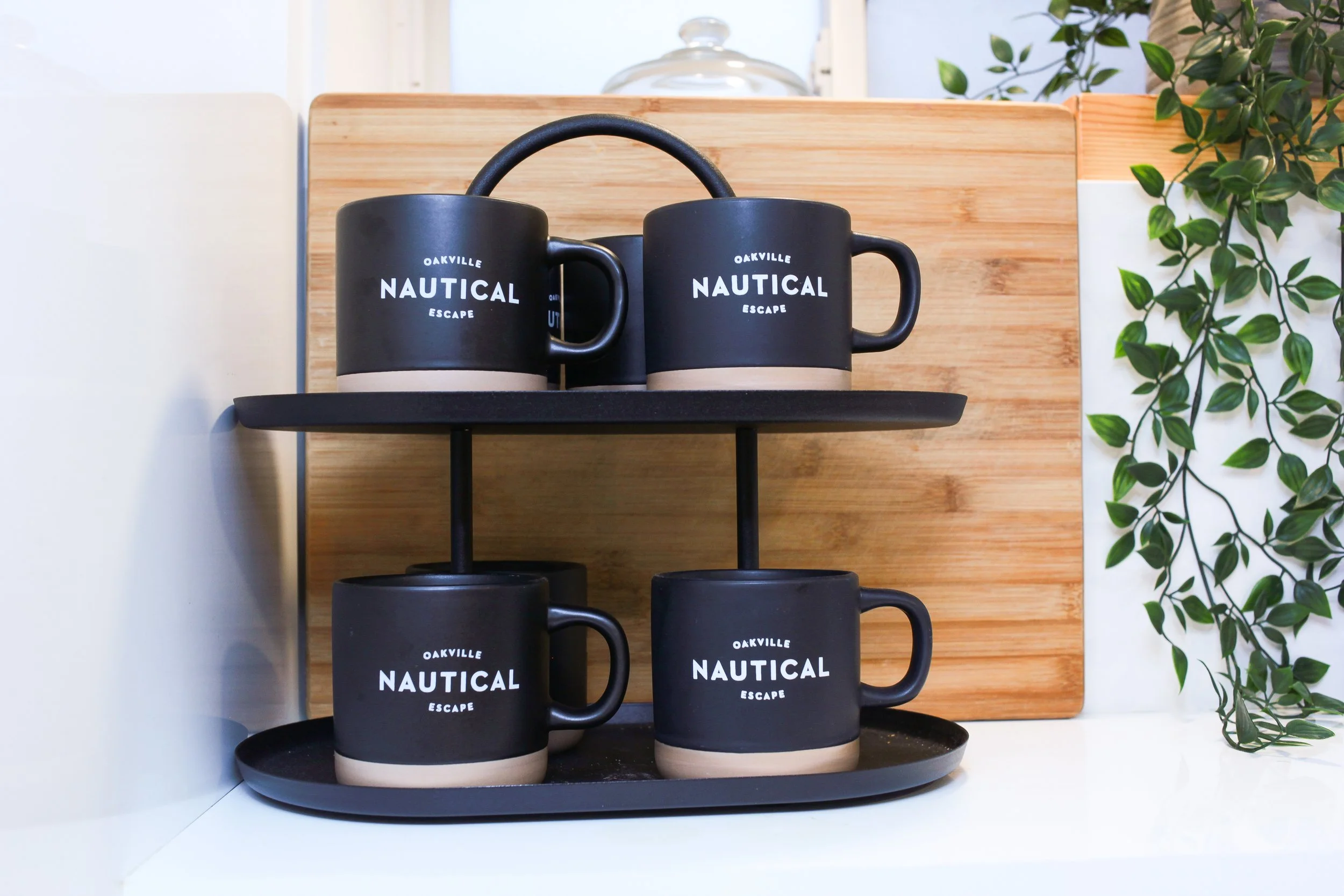 Four black coffee mugs with beige bases, labeled 'Oakville Nautical Escape,' on black tiered serving trays, with a wooden cutting board and green leafy plant in the background.