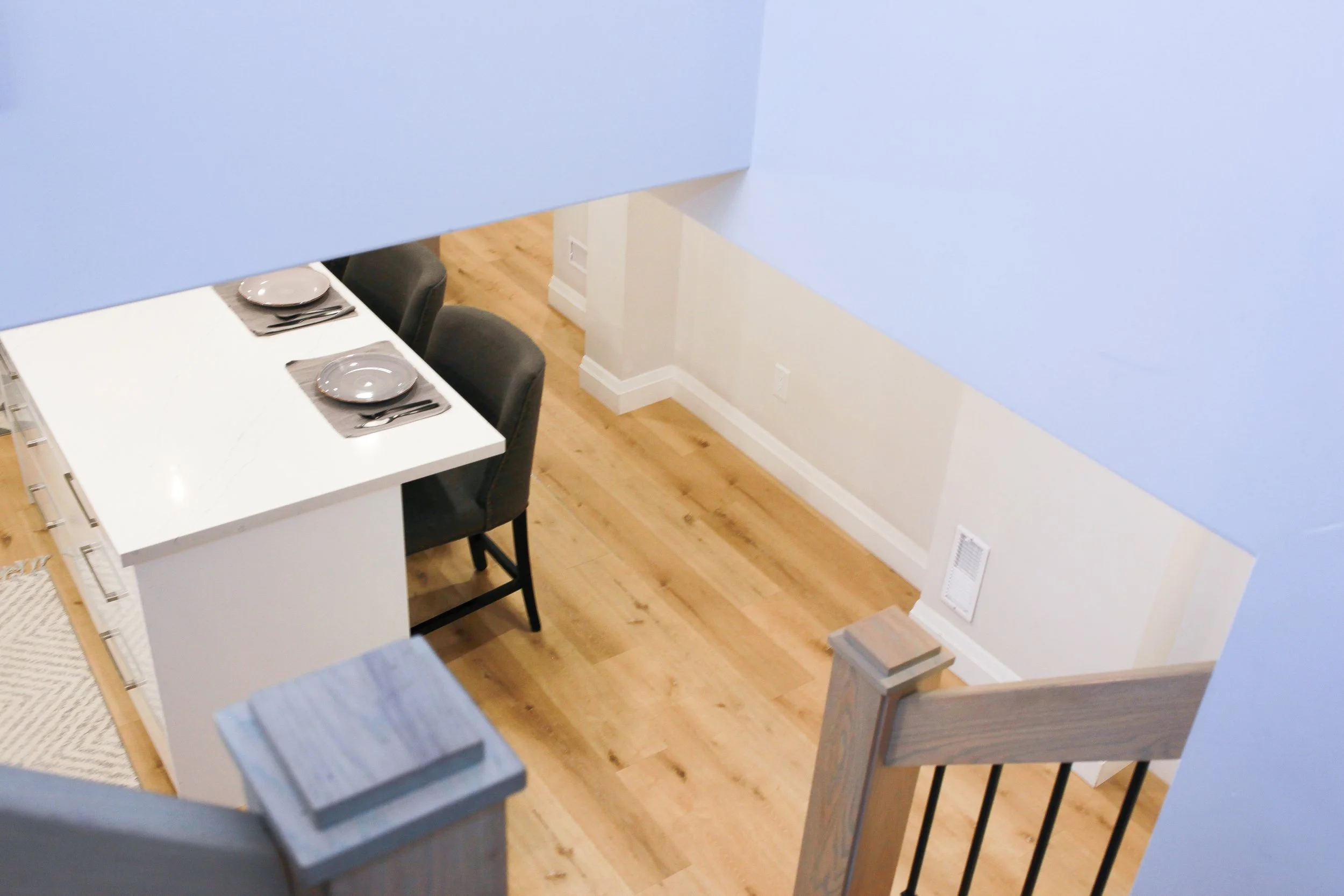 Overhead view of a dining area with a white table, three gray chairs, set with gray plates and napkins, light wood flooring, and white walls.