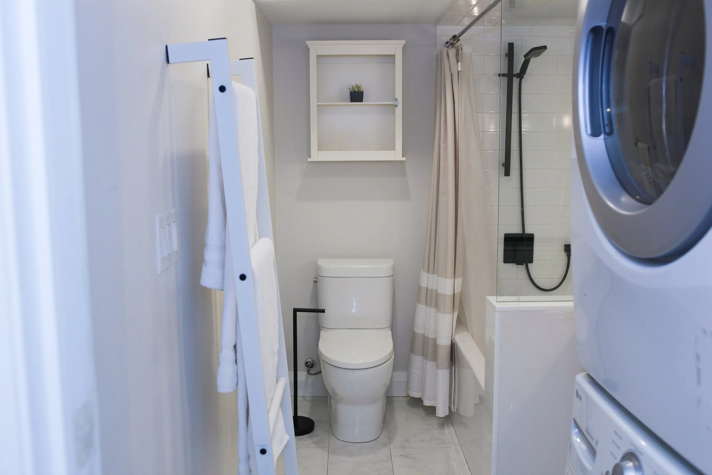 A compact bathroom with a white toilet, a beige shower curtain, a black showerhead, and white tiled walls. There is a small white shelf with a potted plant, a laundry ladder with towels, and a stack of a washing machine and dryer.