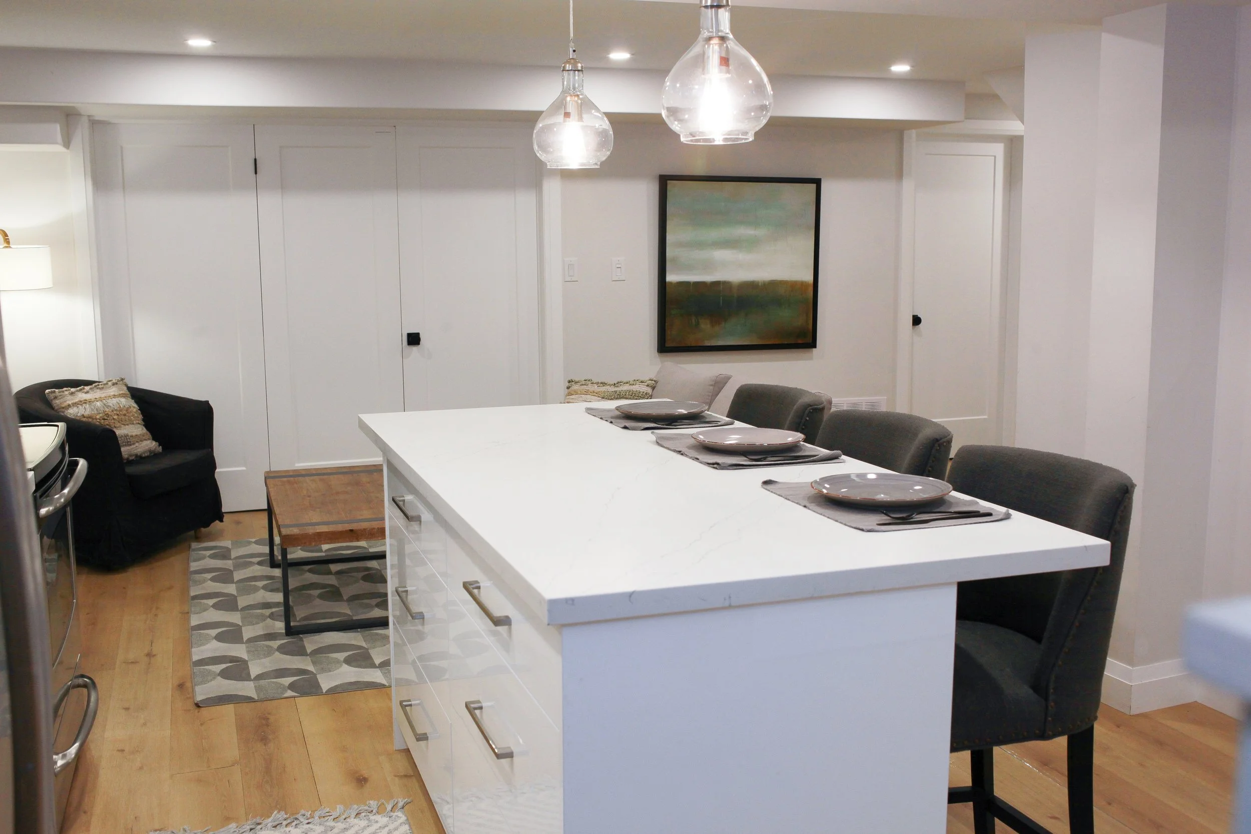 Modern kitchen with white island counter, four chairs, and a white door, with artwork on the wall and pendant lights overhead.