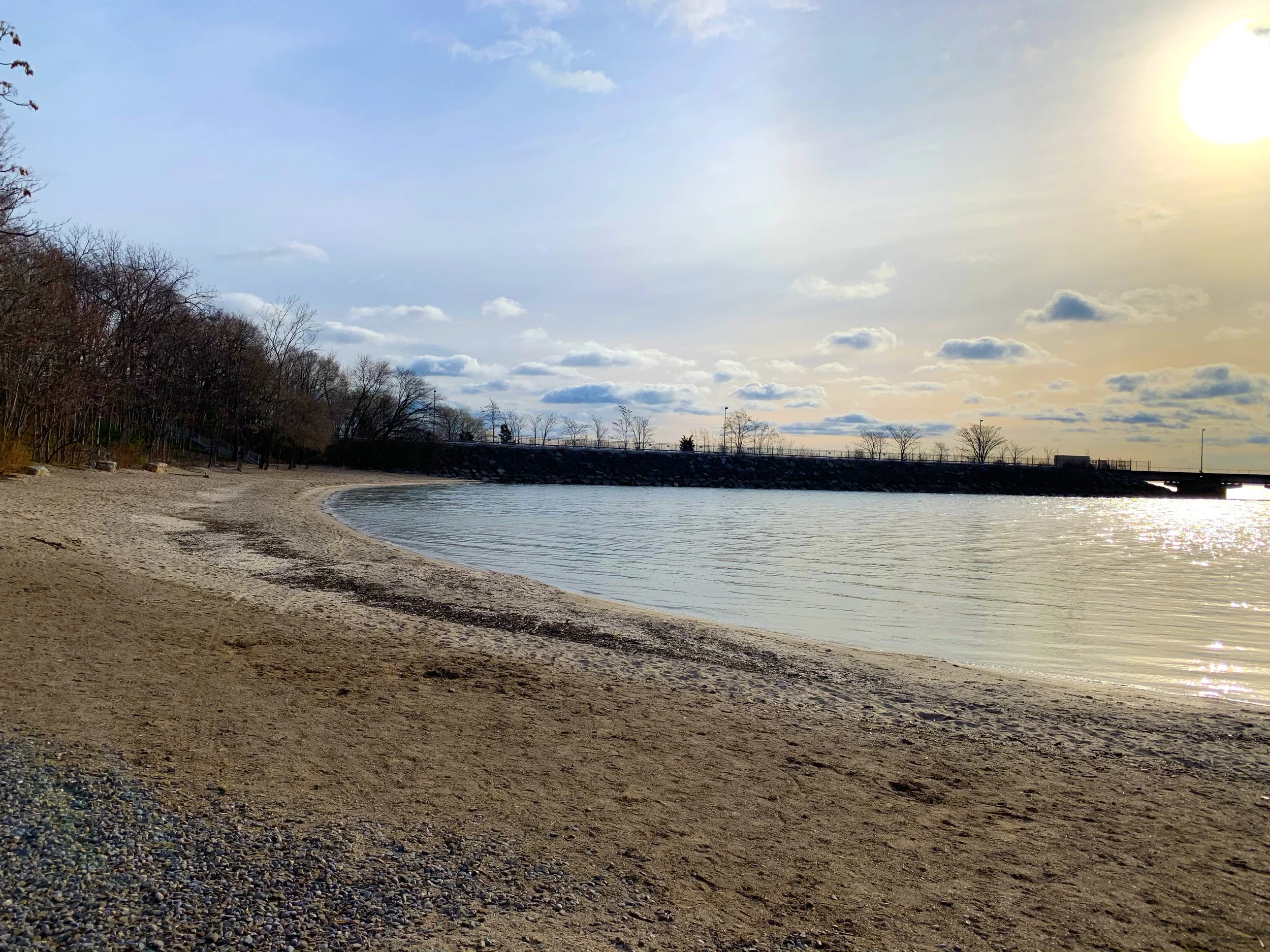 A sandy beach along a calm body of water with a stone wall and trees in the background. The sky is partly cloudy with the sun shining.