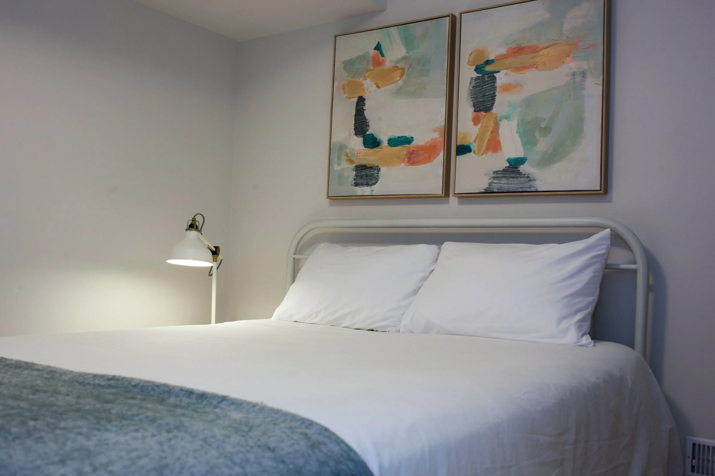 A bedroom with a white bedspread, pillows, a bedside lamp, and two abstract art paintings on the wall.