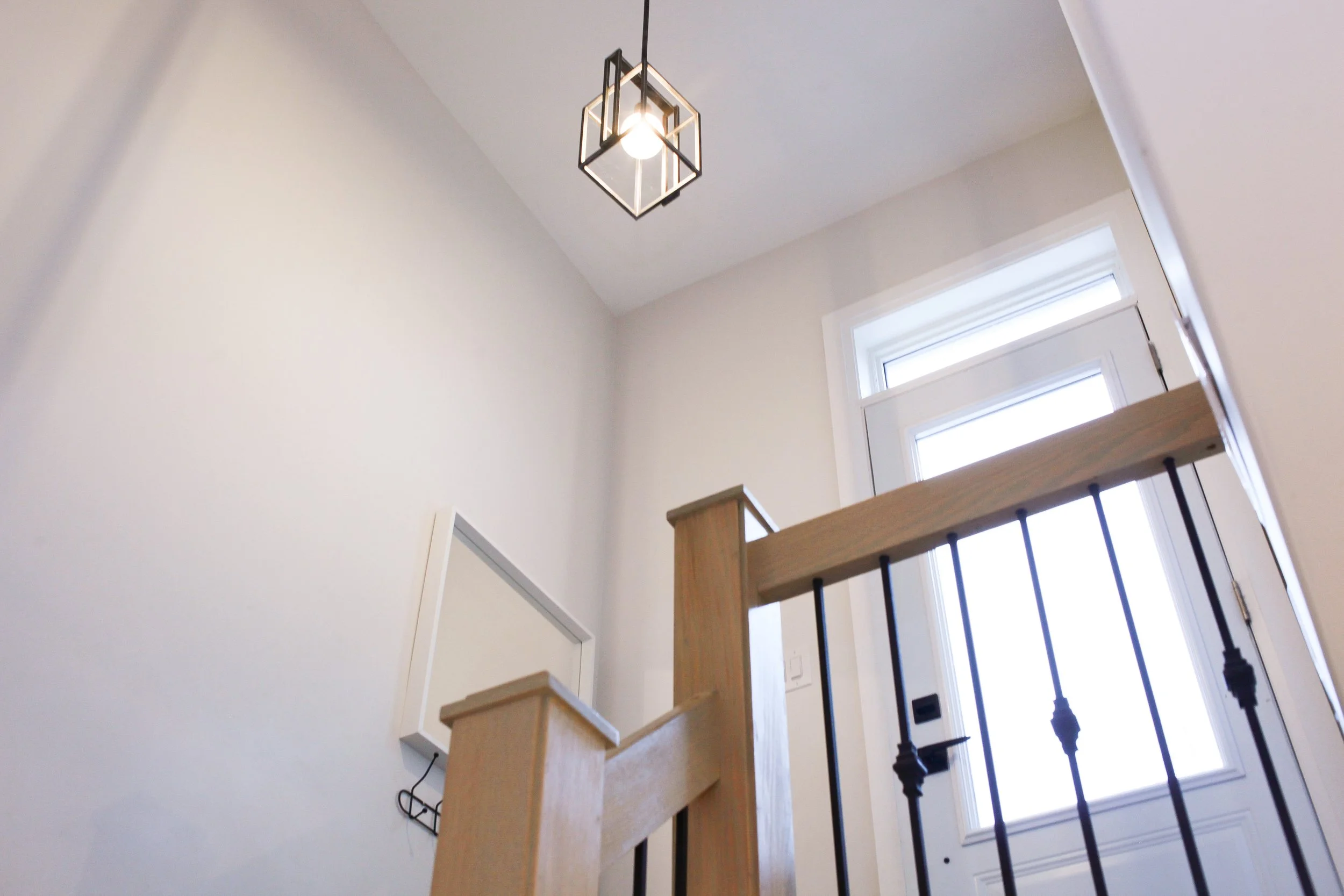 View of a house's interior staircase with wooden handrail and black metal balusters, a white wall, and a window near the door, with a modern geometric ceiling light fixture.