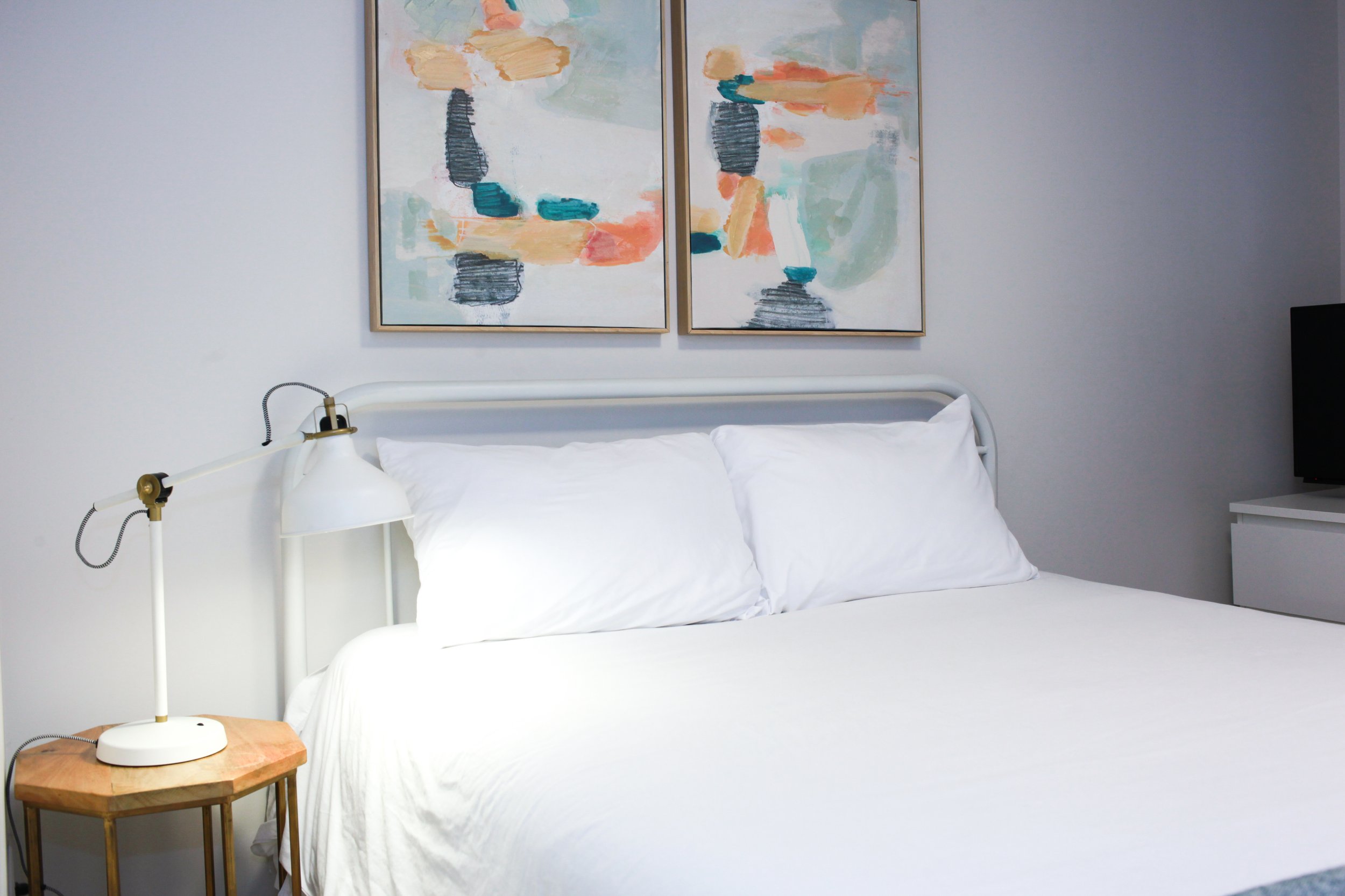 A neatly made bed with white sheets and pillows, a white headboard, and a wooden nightstand with a white lamp. Two abstract paintings with pastel and dark colors hang above the bed, with a television on a white dresser to the right.