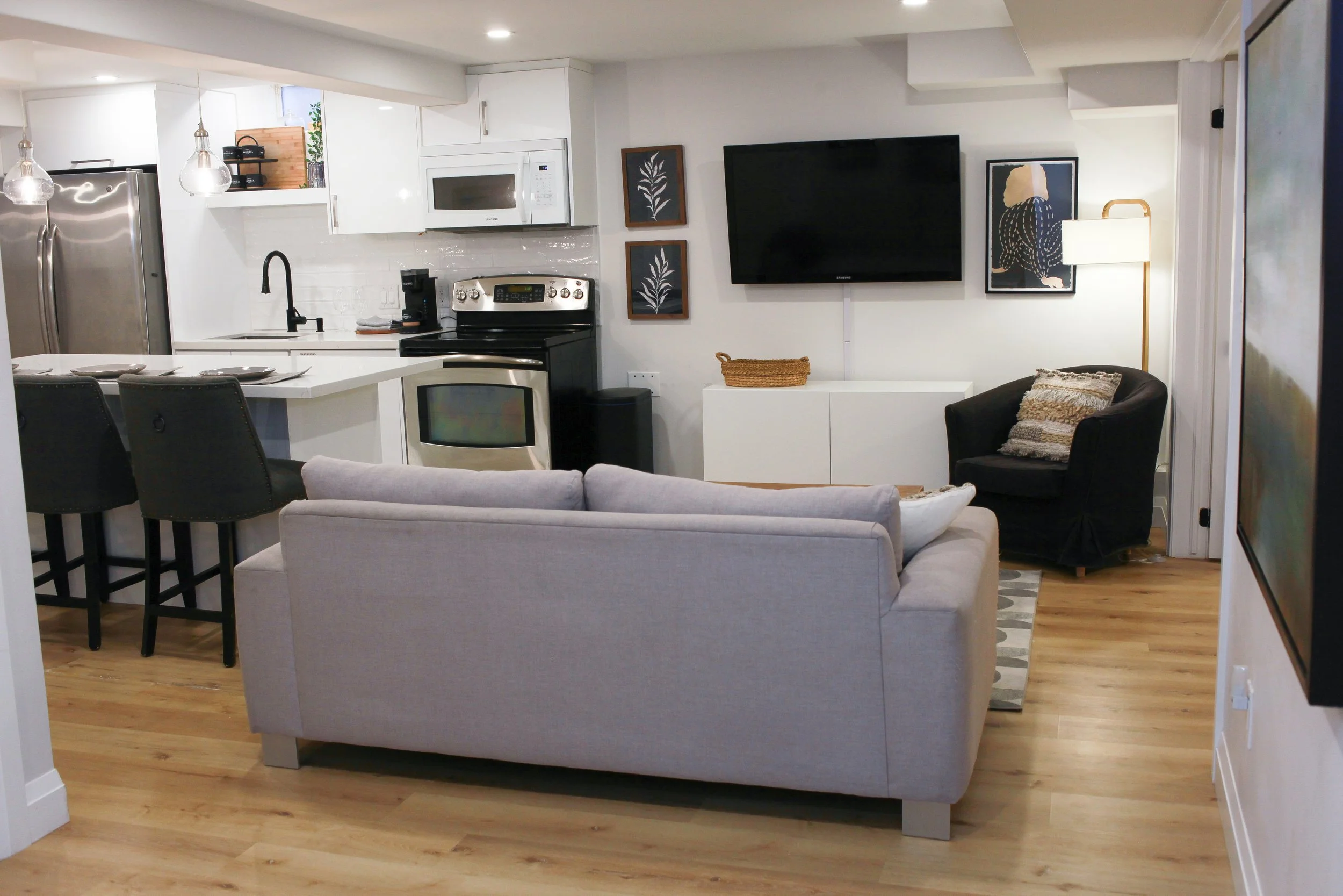 Open-concept living room and kitchen with a light gray sofa, black armchair, mounted flat-screen TV, white cabinetry, stainless steel appliances, bar-style seating with three black chairs, and modern decor.