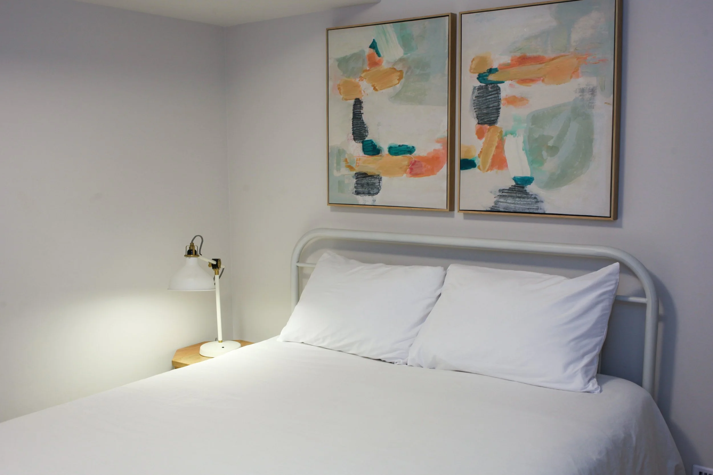 A neatly made bed with white pillows and bedding, a small wooden side table, and a modern white and gold desk lamp. Above the bed, there are two abstract paintings with pastel colors on a white wall.