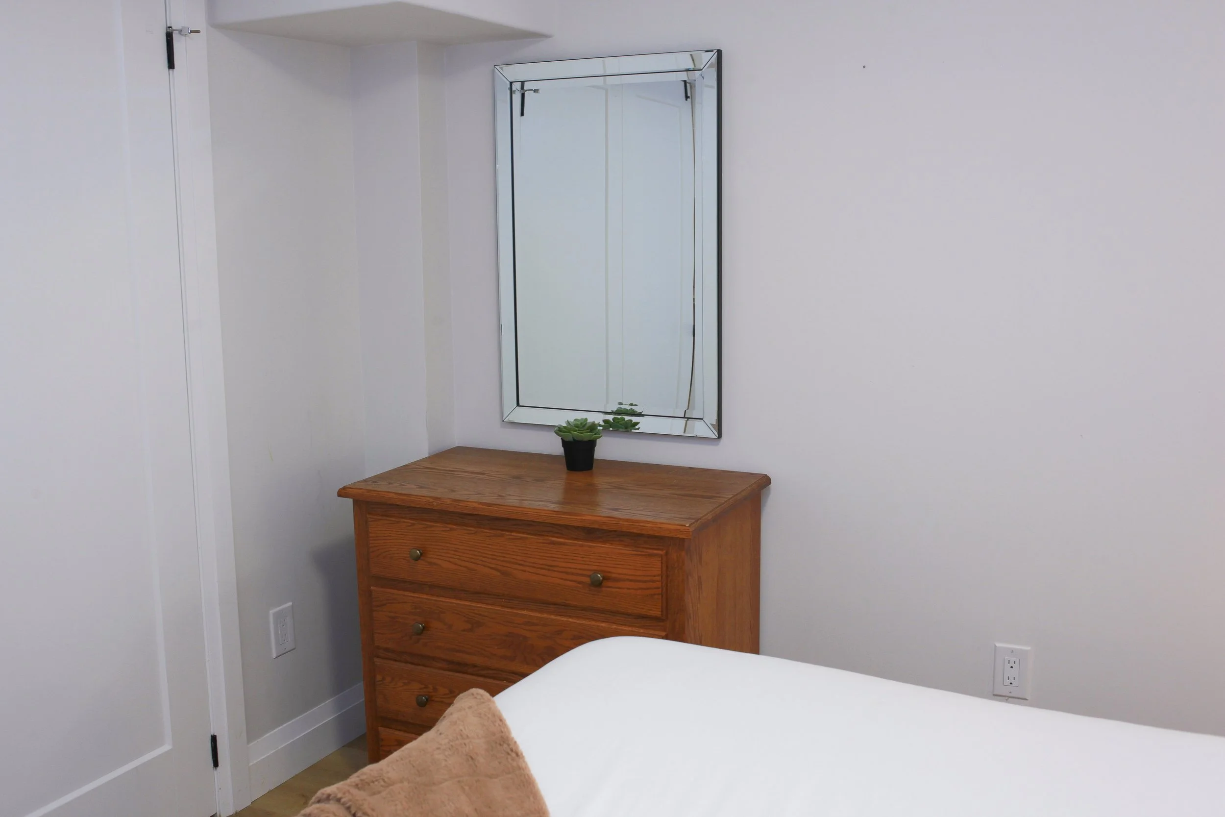 A bedroom corner with a wooden dresser, a mirror above it, a small potted plant, and part of a bed with a brown blanket.