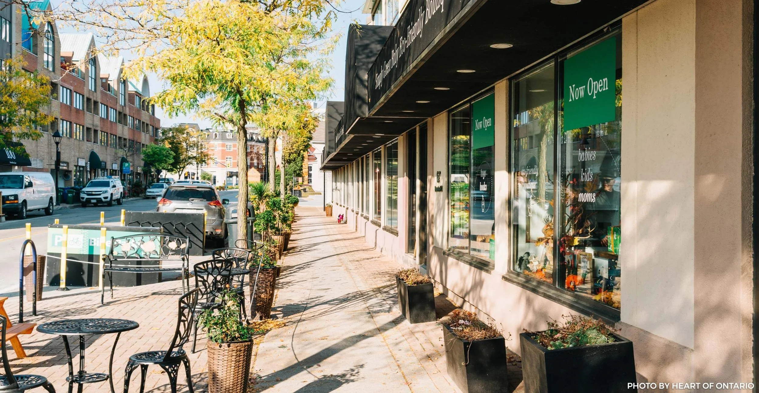 Street view of a sidewalk outside a store with green 'Now Open' signs in the windows, outdoor seating, potted plants, and cars parked along the street.