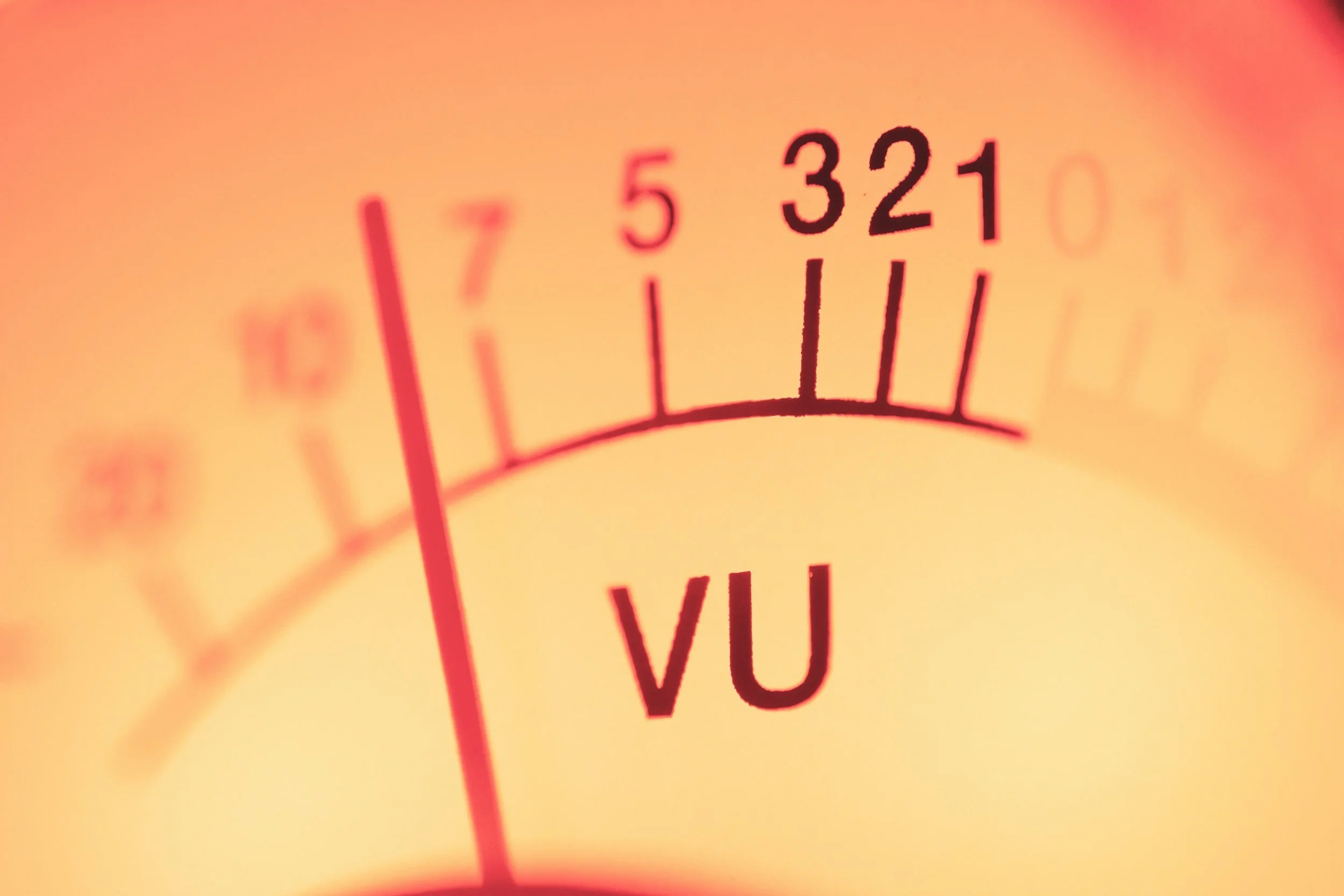 Close-up of a VU meter with a red needle pointing to the 7 mark, used for measuring audio signal levels, with a warm orange background.