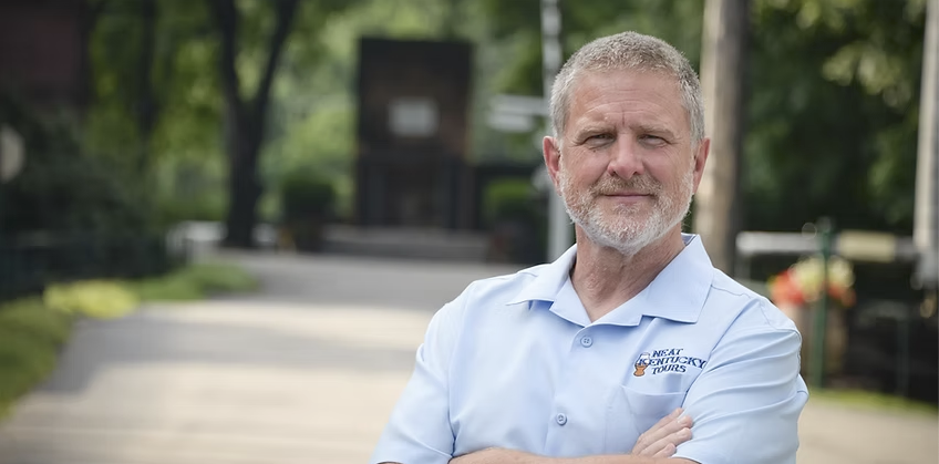 A middle-aged man with gray hair and beard standing outdoors with arms crossed, wearing a light blue collared shirt with a logo that reads 'Great Kentucky Tours'.
