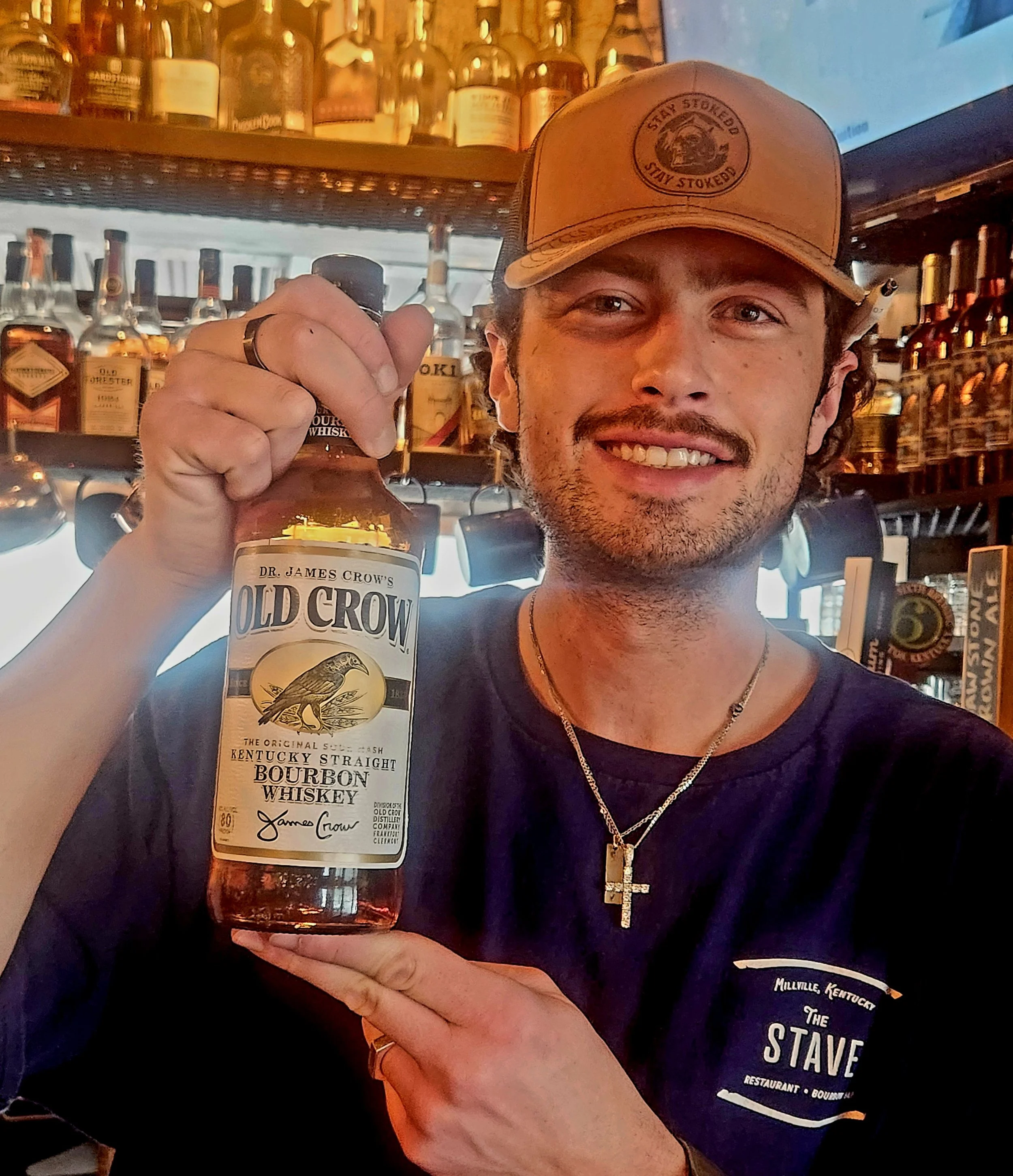 A man wearing a tan baseball cap and black shirt holding a bottle of Old Crow Kentucky Straight Bourbon Whiskey inside a bar.