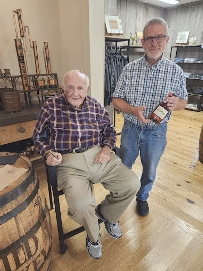 Two men, one sitting and one standing, inside a store with wooden floors and decor. The seated man is elderly, wearing a plaid shirt and beige pants. The standing man is middle-aged or older, wearing a plaid shirt and jeans, holding a bottle.