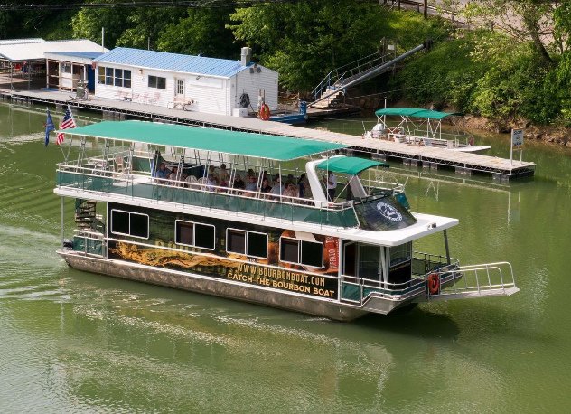 A two-story boat with a green roof moving through a river. A sign on the side promotes bourbon, and there are houses and docks along the riverbank.