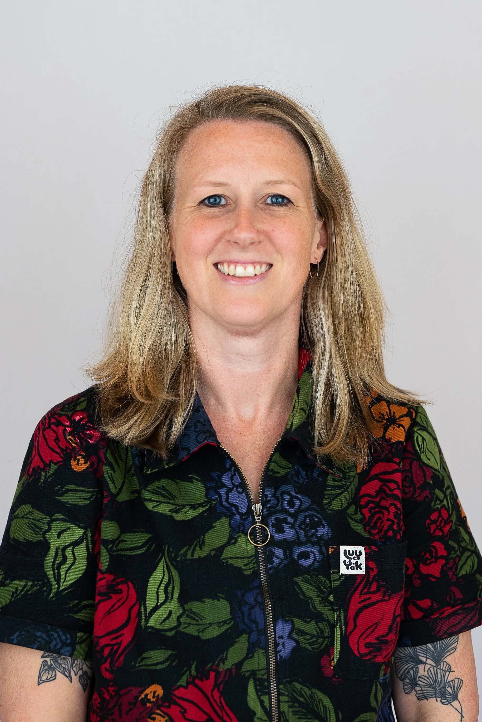 A woman with shoulder-length blonde hair and blue eyes smiling, wearing a black floral patterned zip-up shirt with red, green, orange, and blue colors.