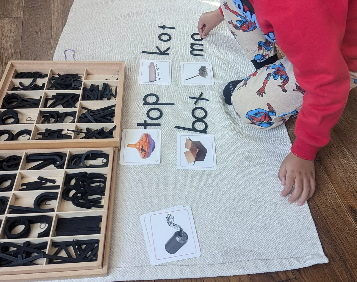 Child playing a word-matching game with picture and letter cards on a mat, with wooden alphabet letter trays beside.