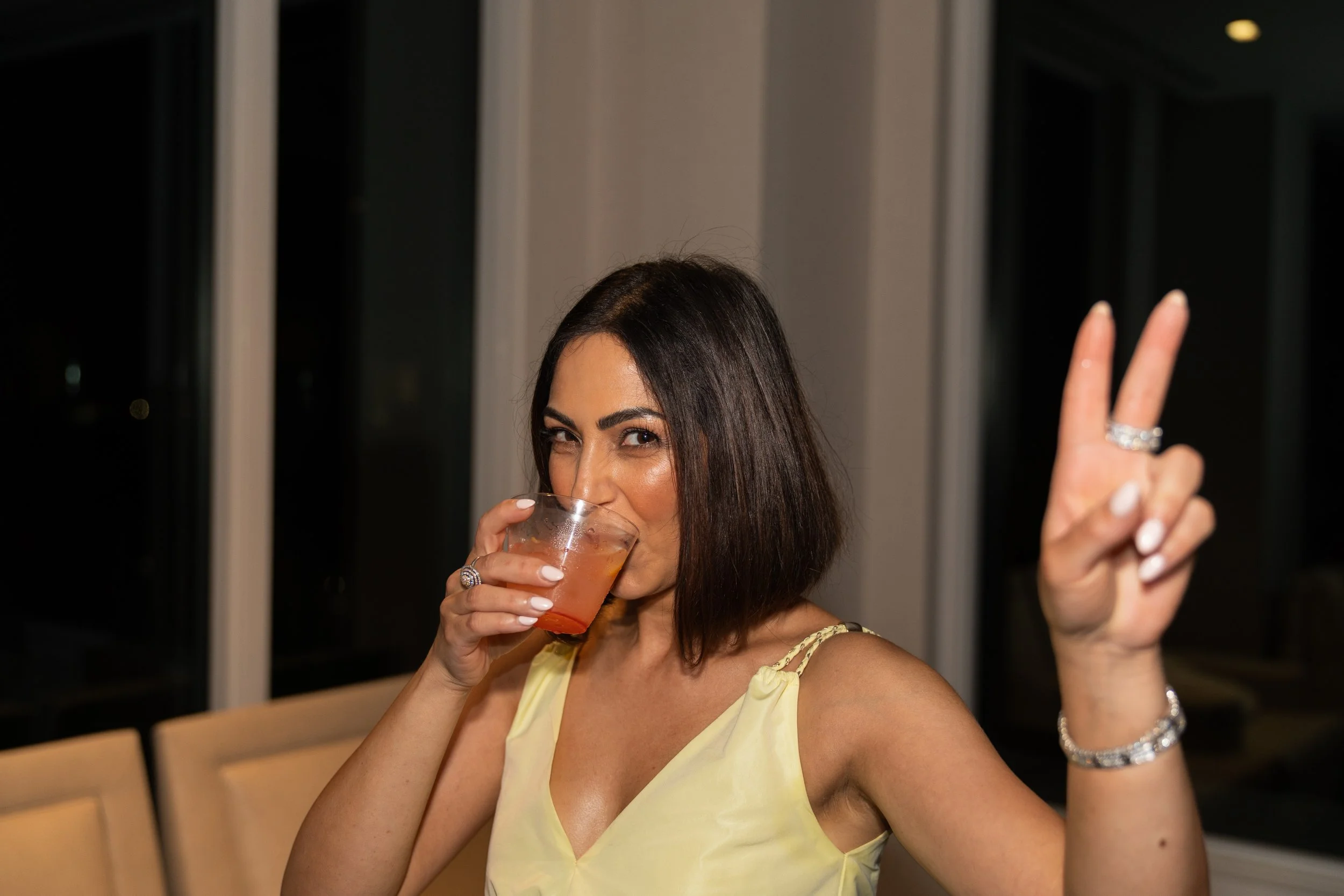 A woman with dark hair, wearing a yellow dress, is holding a glass with a drink in one hand and making a peace sign with the other hand. She is smiling and appears to be at a party or celebration.