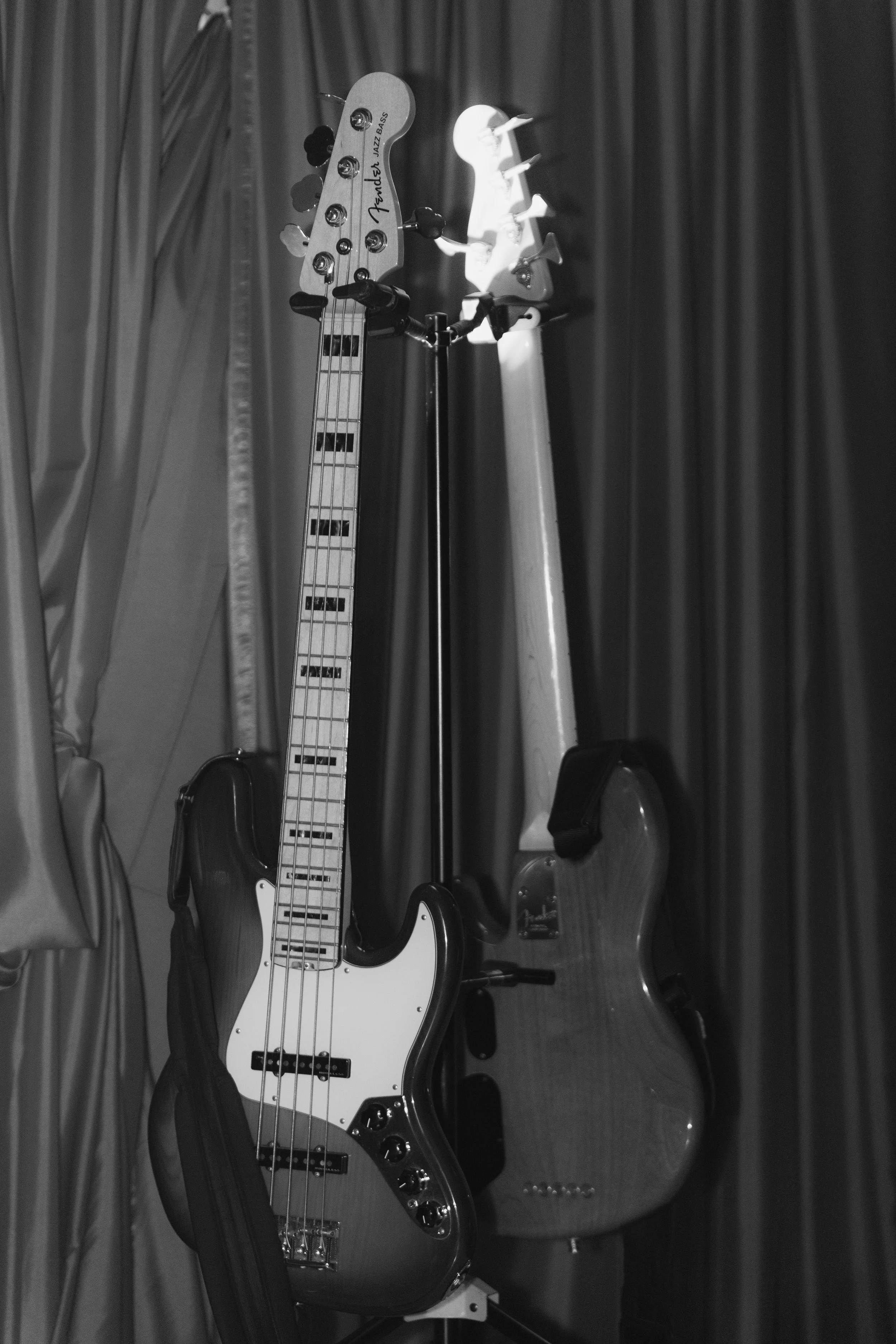 Two electric guitars, one Fender Jazz Bass and one Fender Stratocaster, leaning against a wall with curtains. A cello is also visible behind them.