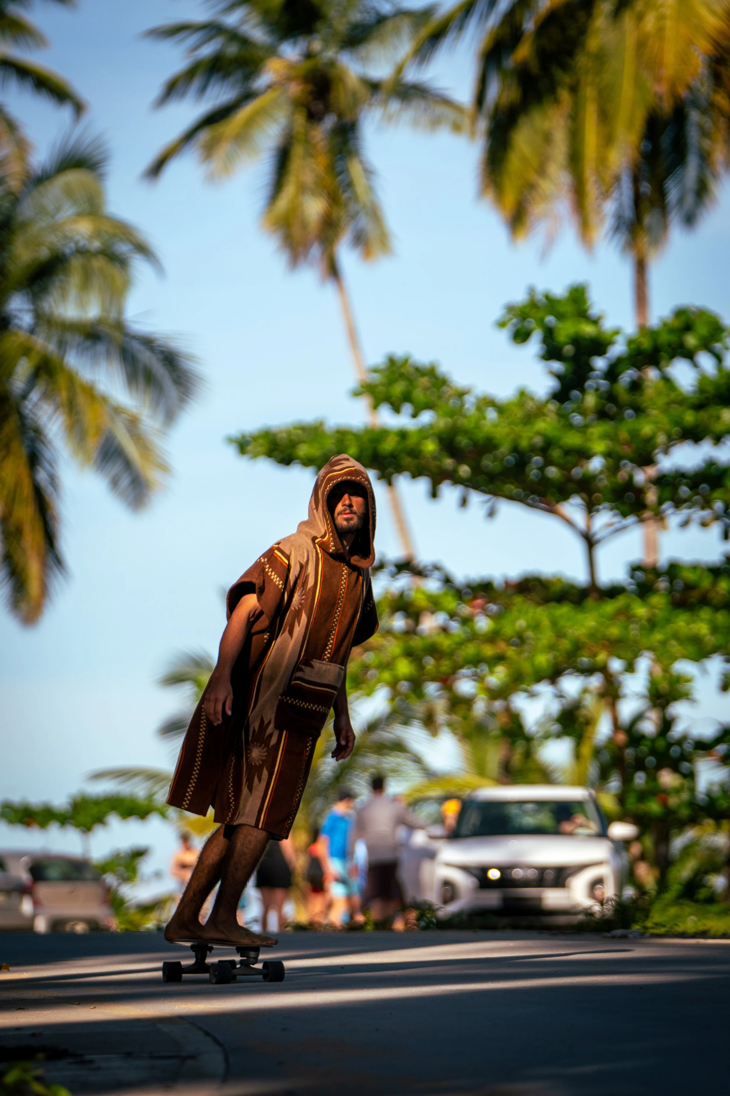 A person dressed in a brown tribal-style hooded cape, riding a skateboard on a street, with palm trees and a light blue sky in the background. Some people and a white car are visible in the blurred background.