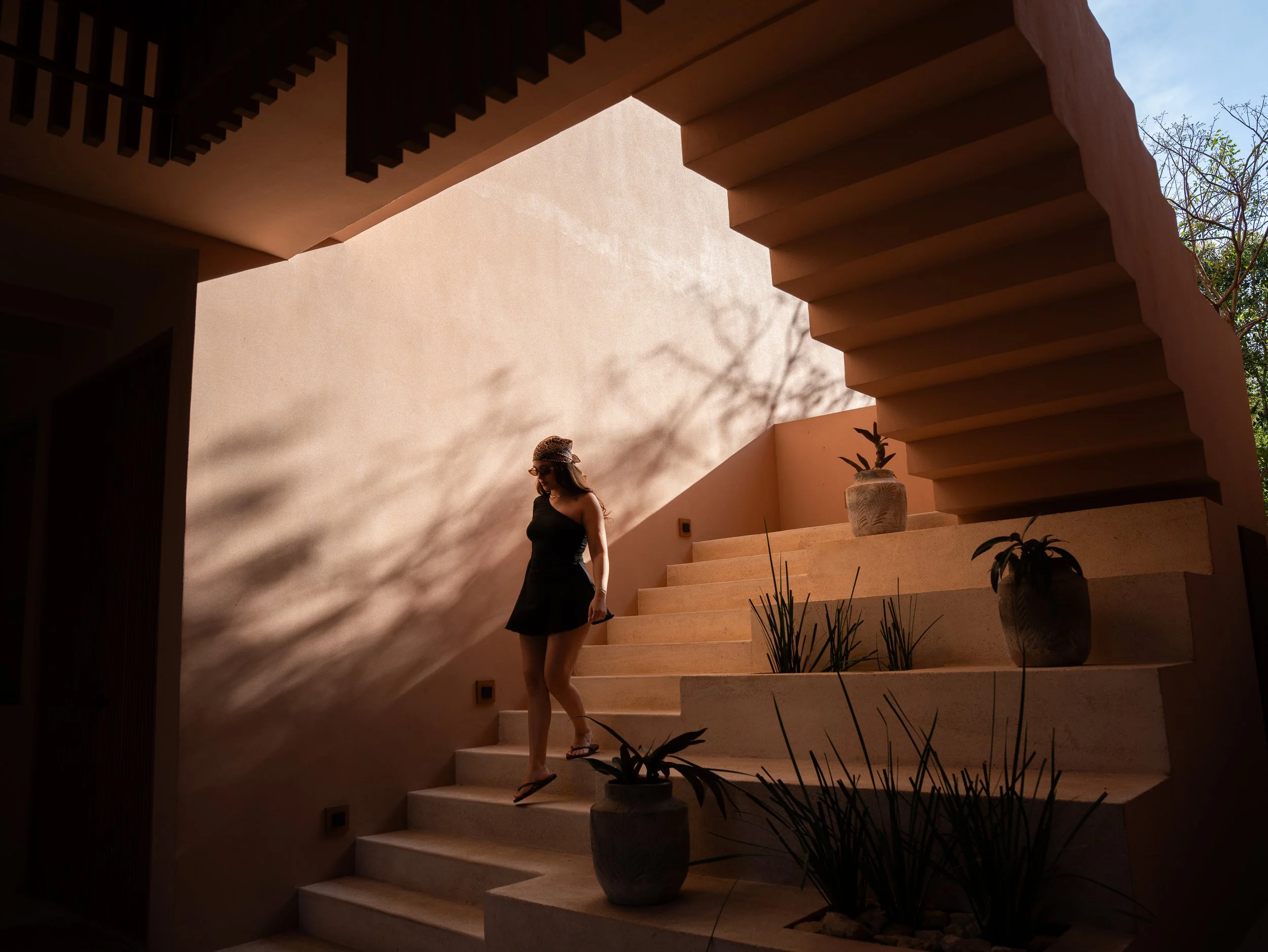 A woman wearing sunglasses and a black dress walking down beige stairs in a courtyard with potted plants and warm sunlight creating shadows on the wall.