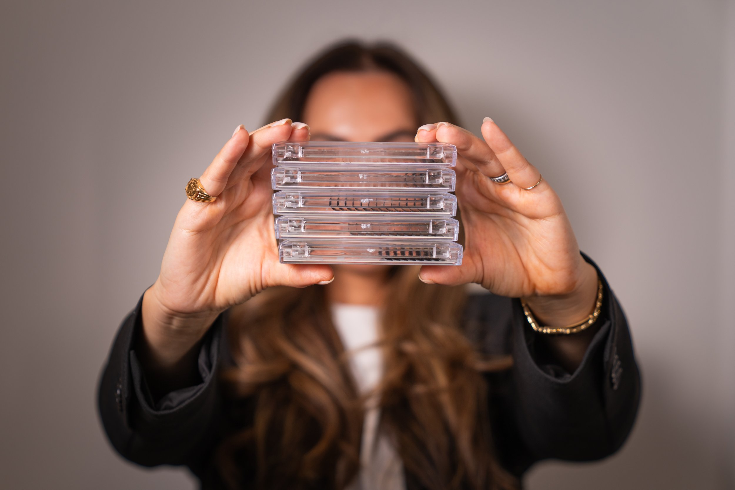 A woman with long brown hair is holding a stack of clear plastic CD cases in front of her face, obscuring her facial features. She is wearing rings on her fingers and a black jacket.
