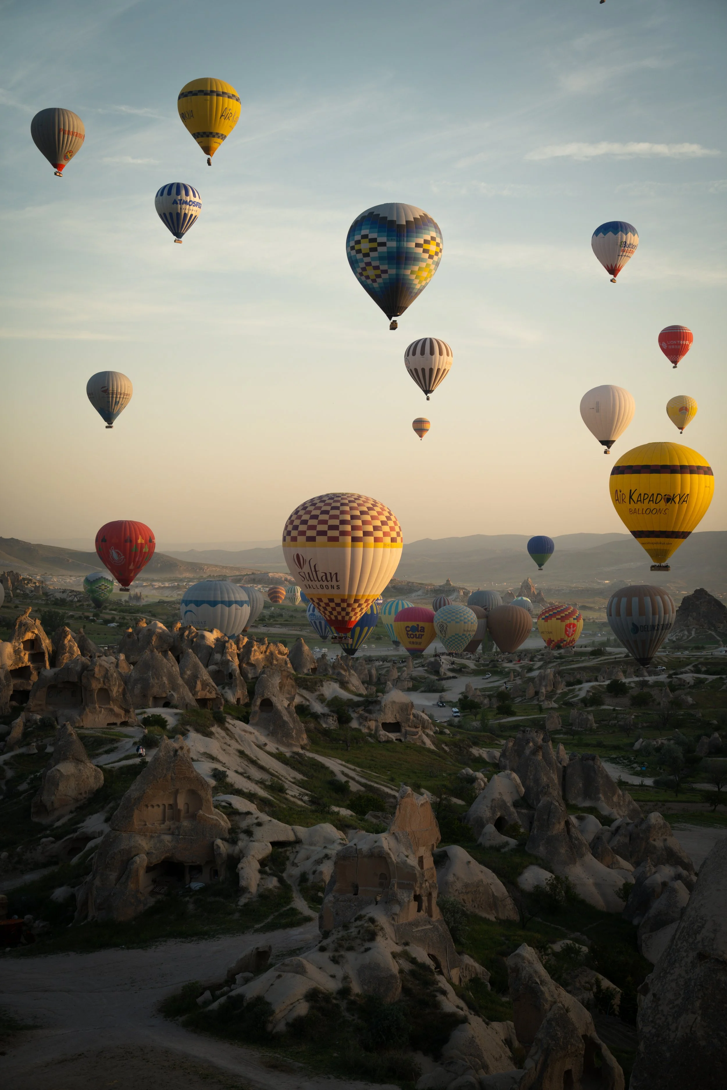 Multiple colourful hot air balloons floating in the sky over a landscape with rocky formations and cave dwellings in Cappadocia, Turkey.