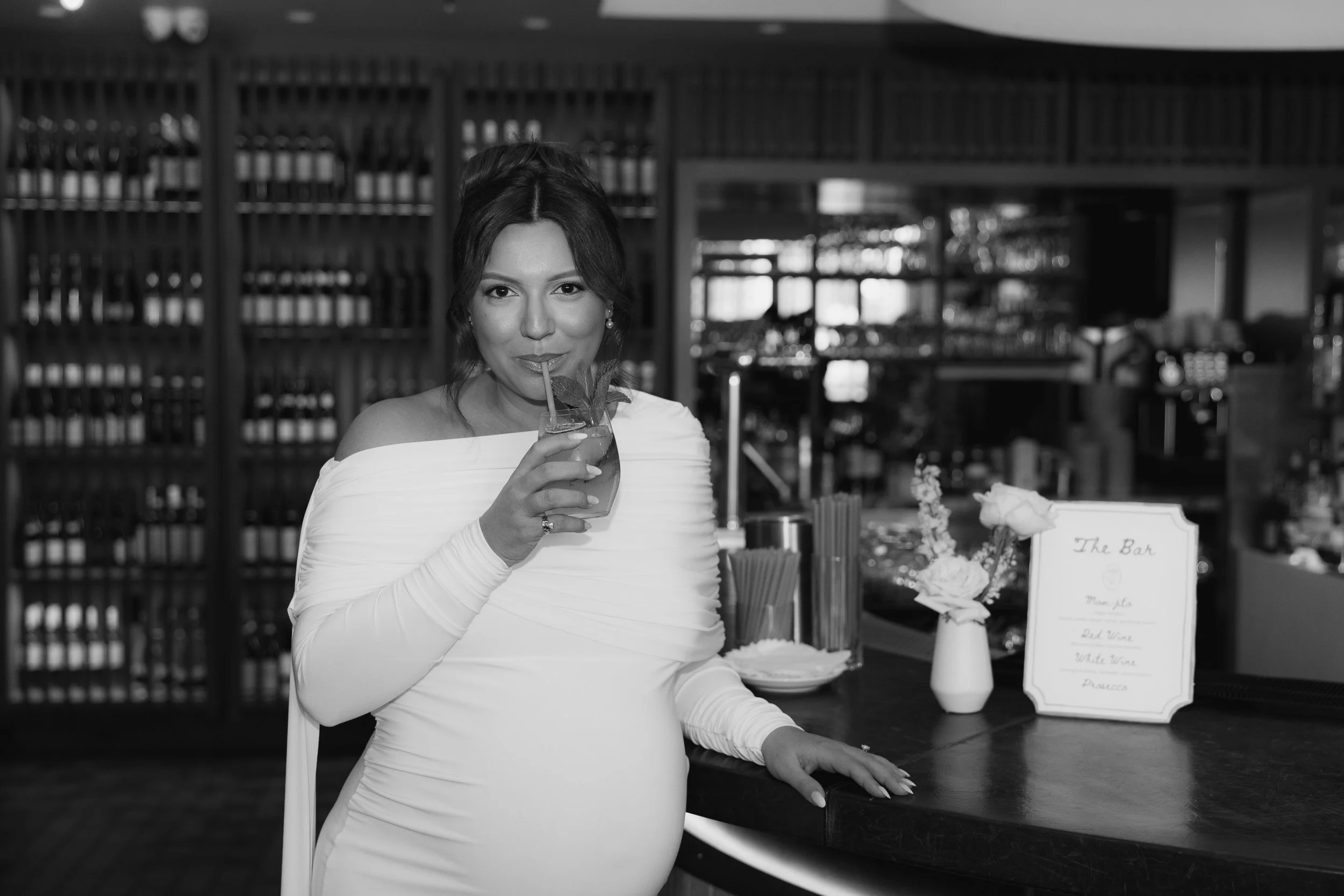 A woman in an elegant off-shoulder dress drinks a cocktail with a straw while standing at a bar with wine bottles in the background. There are flowers and a sign on the bar.