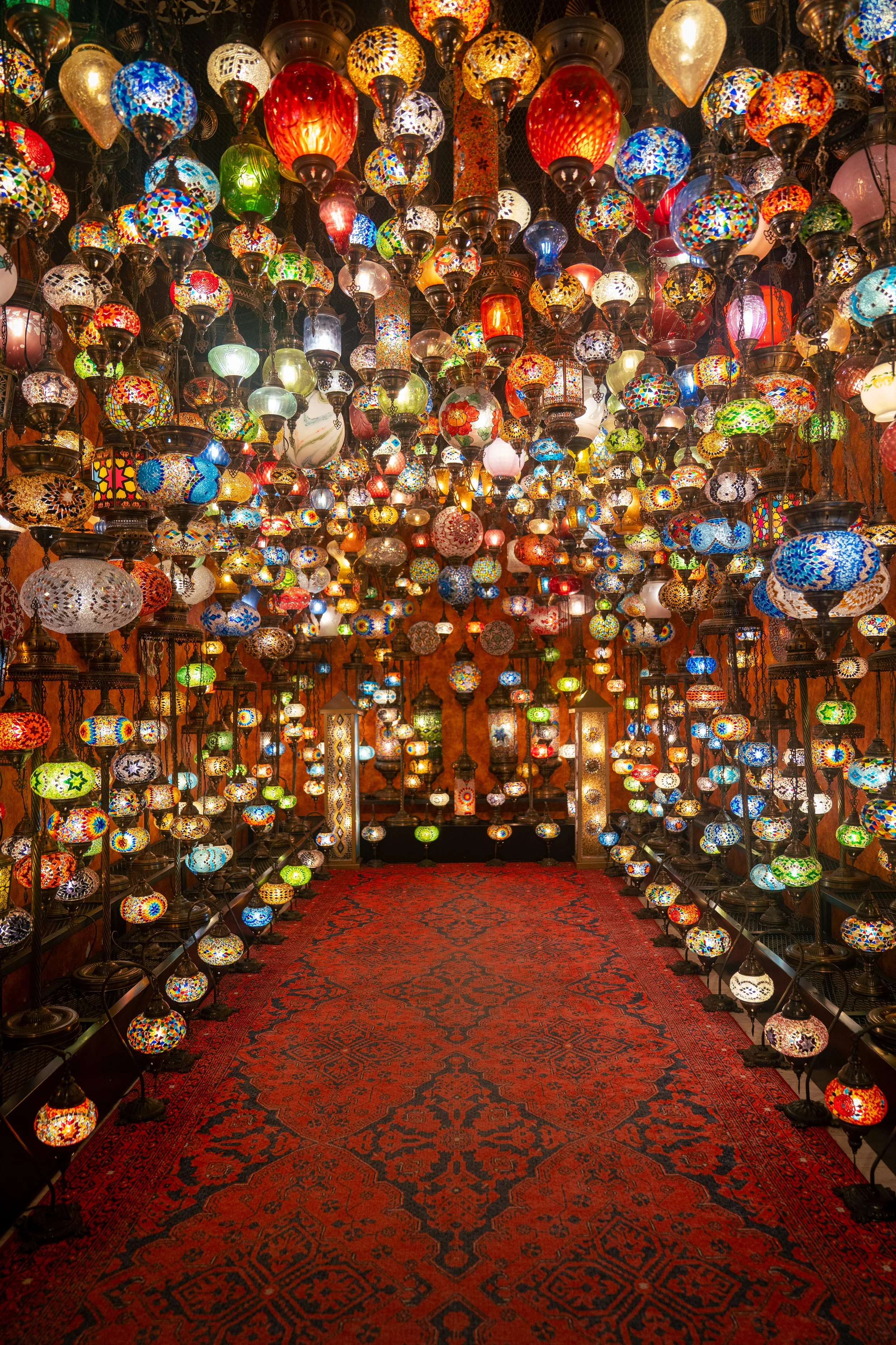 A room filled with hanging colorful mosaic glass lamps and lanterns, with a red oriental rug on the floor.