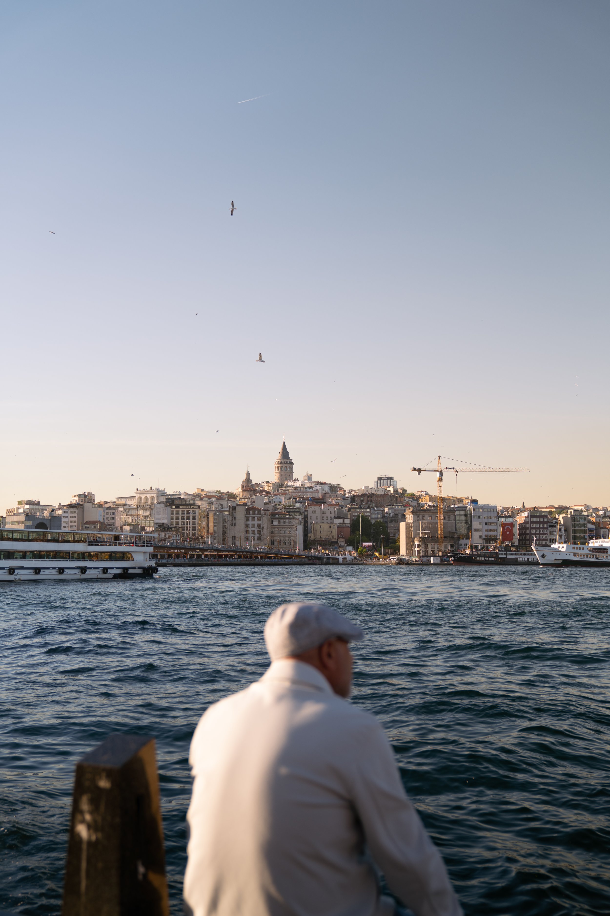 A man in a white outfit and hat sitting on a boat or dock, looking out over the water towards a city skyline with historic and modern buildings, including the Galata Tower, under a clear sky with birds flying.