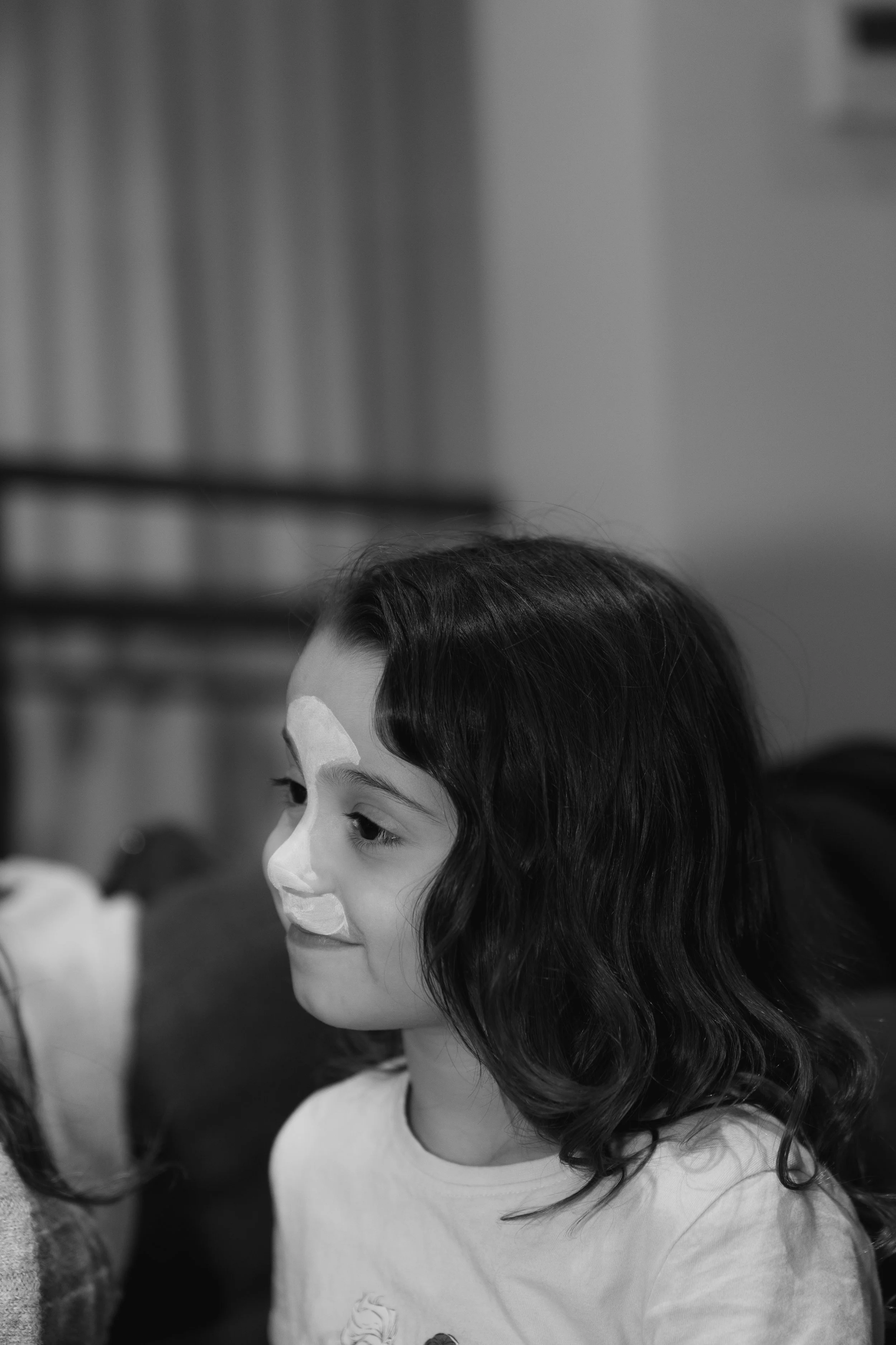 A young girl with long dark wavy hair, smiling and wearing a shirt with a cartoon graphic, sitting in a room with plain walls, possibly at a social or festive gathering.