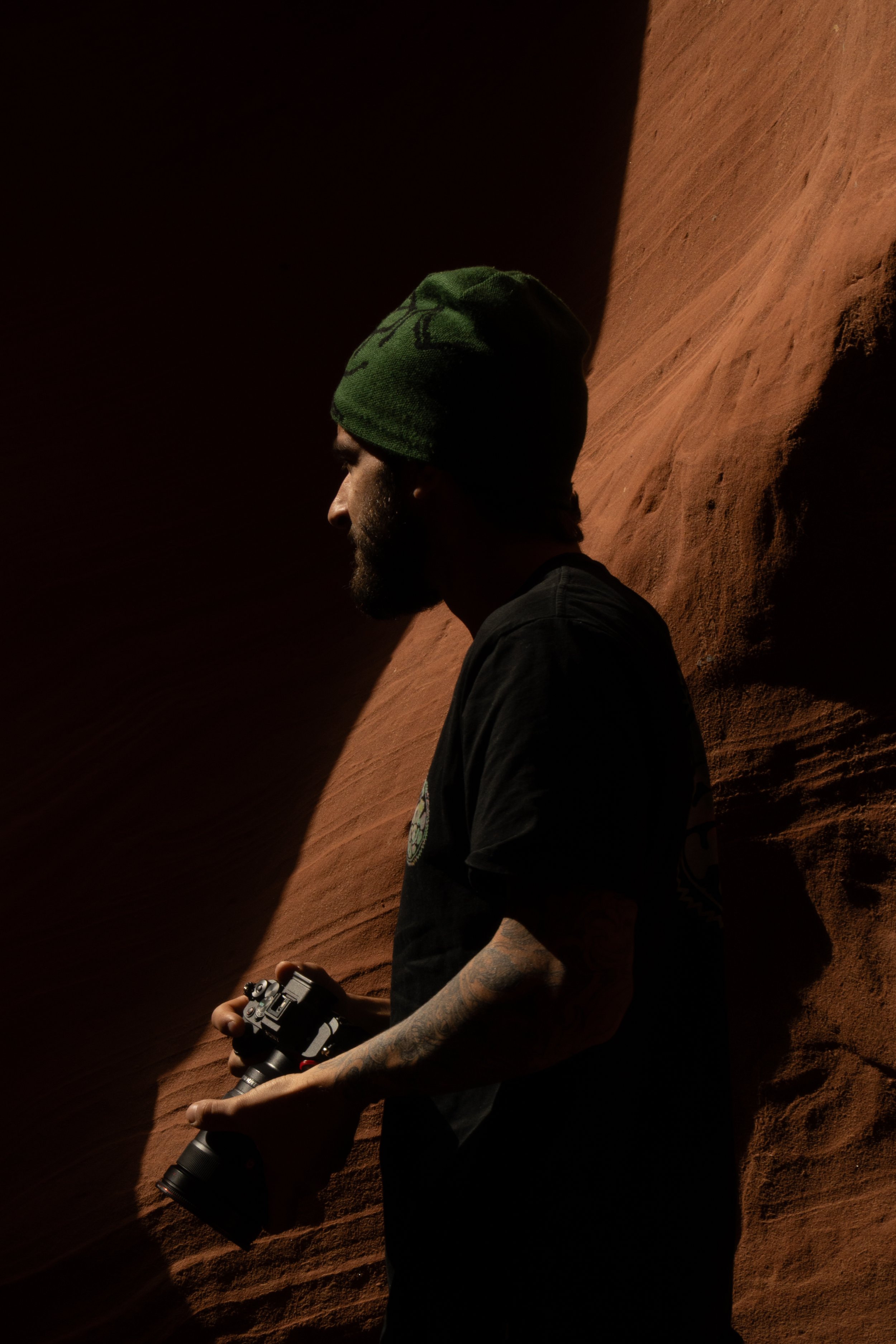 A man with a beard and tattoos on his arm, wearing a green beanie and a black t-shirt, holding a camera in his hands, standing against a red rock formation with sunlight creating shadows.