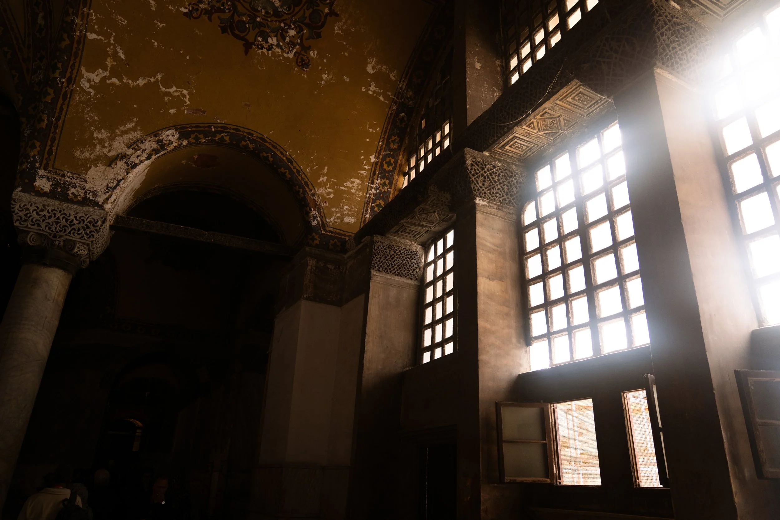 Interior of an old building with large patterned windows, a high arched ceiling with peeling paint, and ornate decorative details on the arches and columns, illuminated by sunlight streaming through the windows.