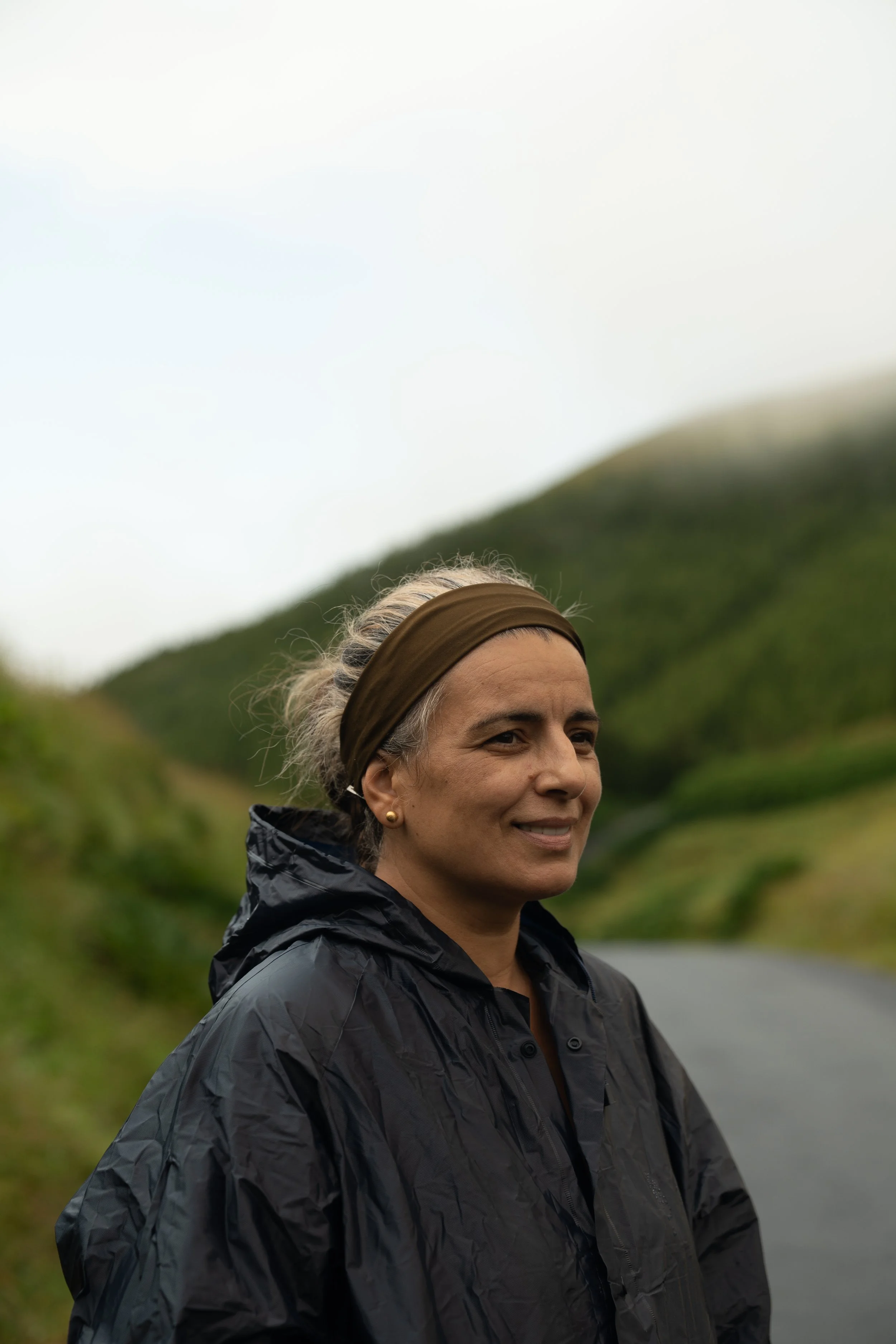 A smiling woman with gray hair tied back, wearing a brown headband and a black waterproof jacket, standing outdoors with green hills in the background under an overcast sky.