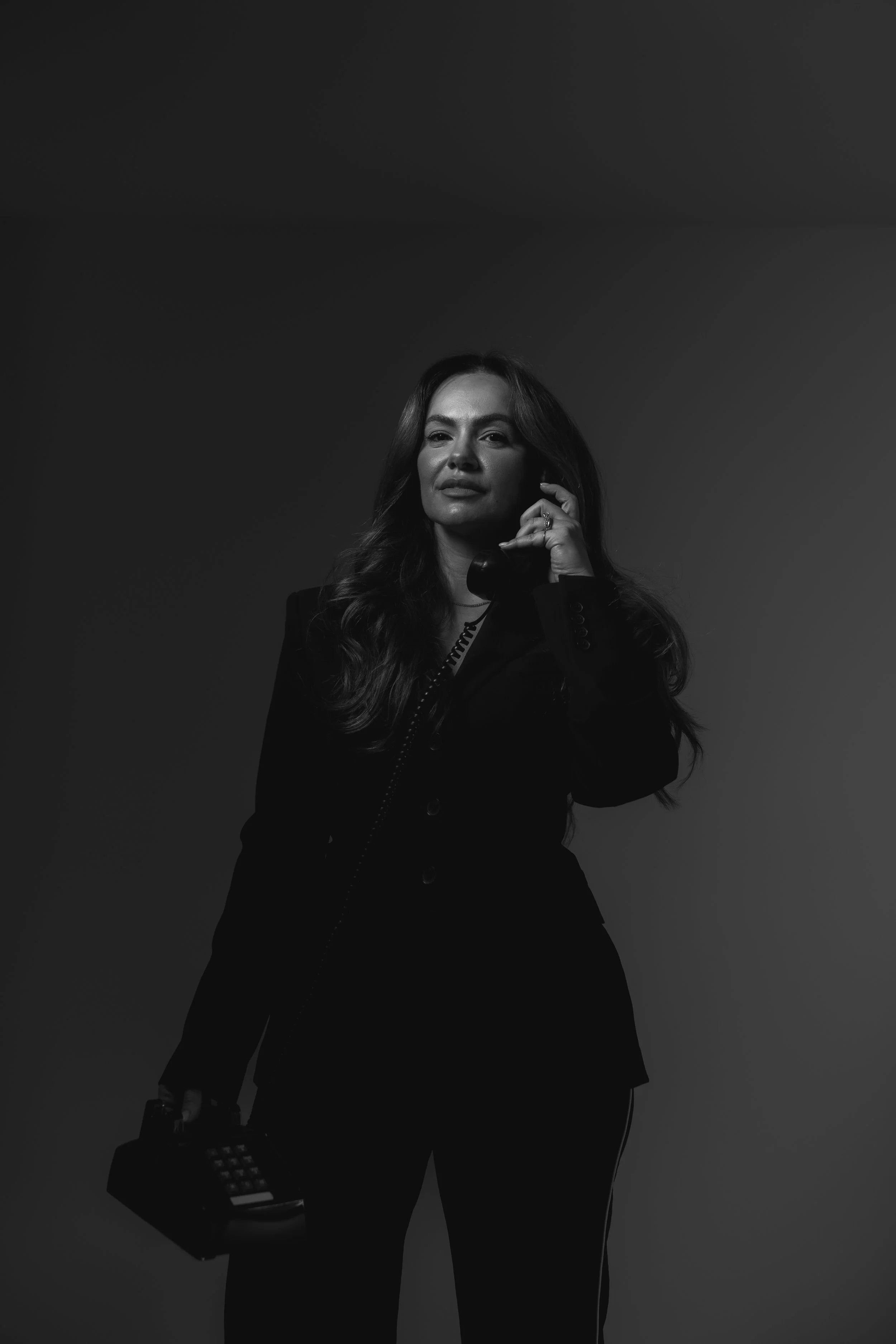A woman with long wavy hair, wearing a dark blazer, is holding a telephone receiver to her ear with her right hand and a vintage calculator in her left hand. She is posing in front of a plain dark background.