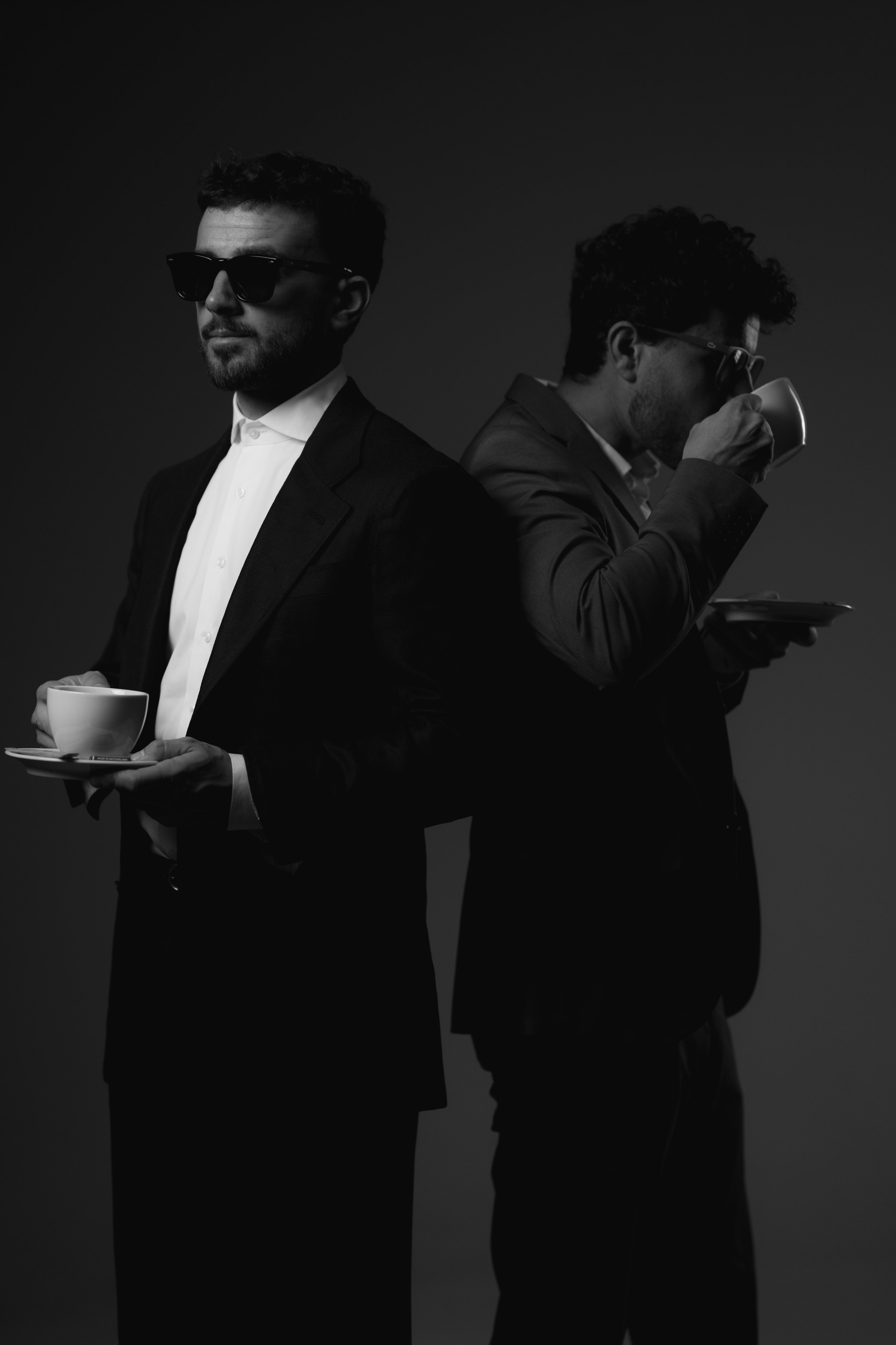 Two men in formal suits, one wearing sunglasses, are standing back-to-back. One is holding a cup and saucer, while the other is drinking from a cup.