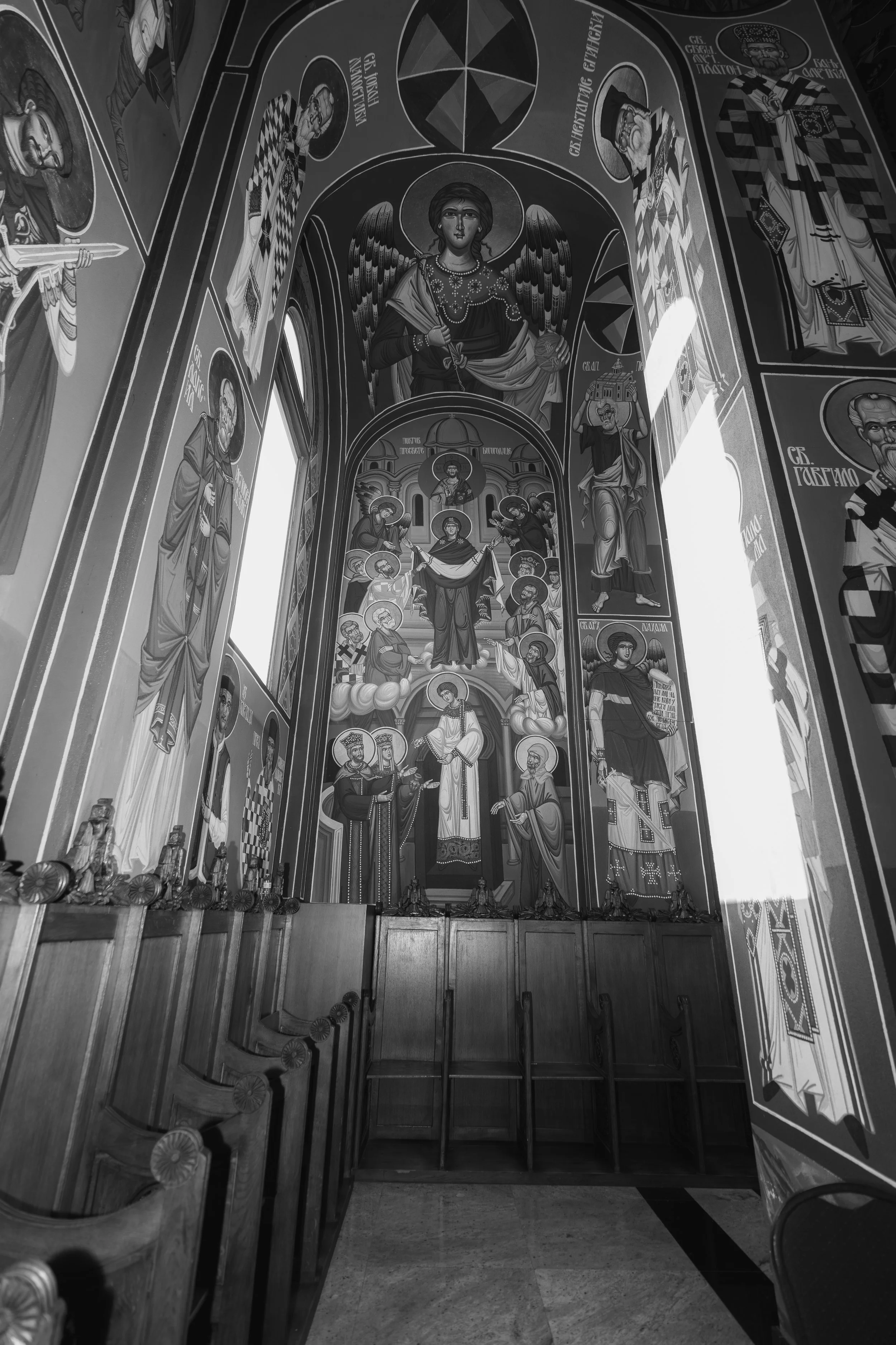 Interior of a church with religious iconography and artwork on the walls and ceiling, including images of saints and angels, with wooden pews and a drawer unit at the bottom.