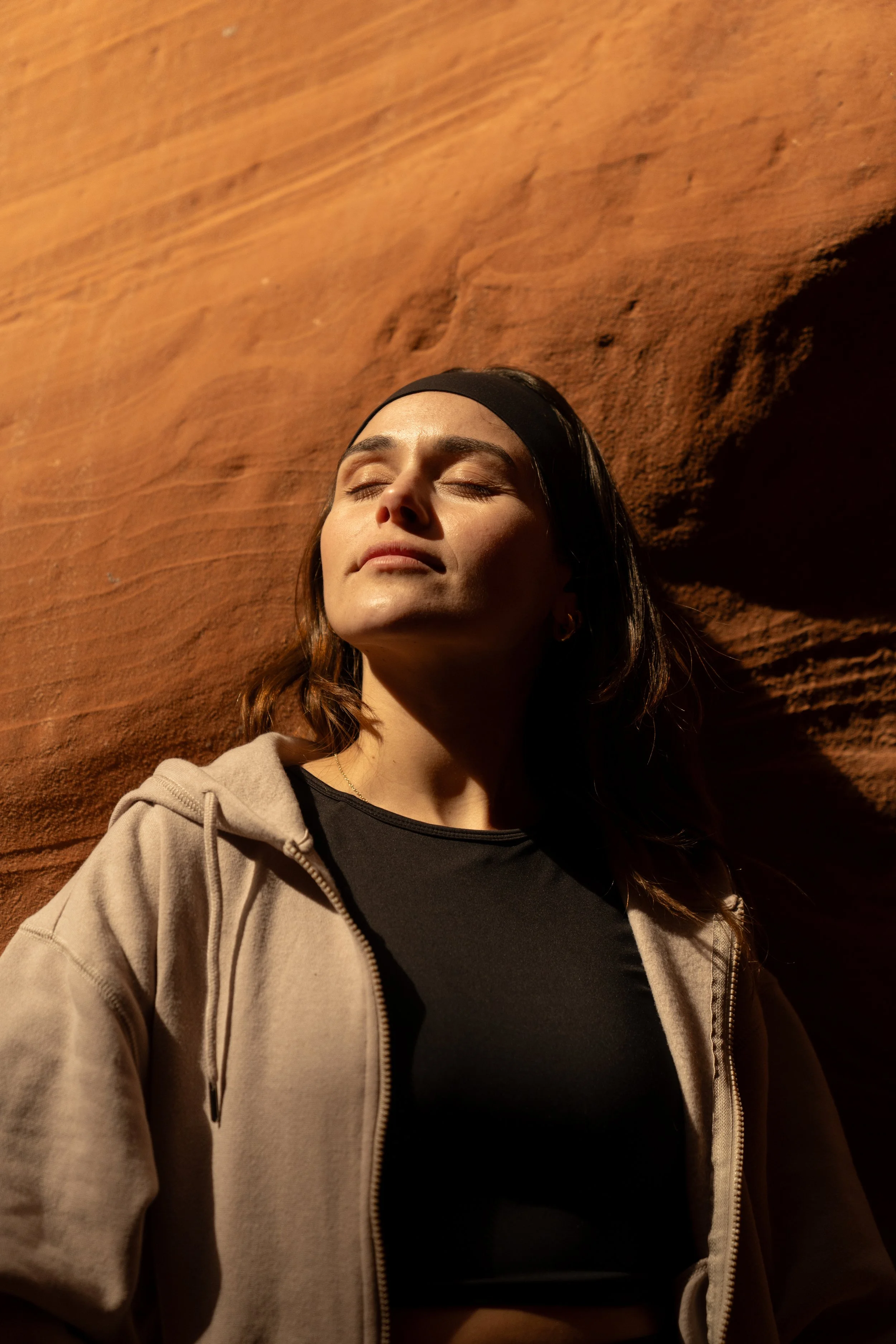Close-up of a woman with dark hair and a headband, her eyes closed, standing against a textured, reddish-brown wall in sunlight.