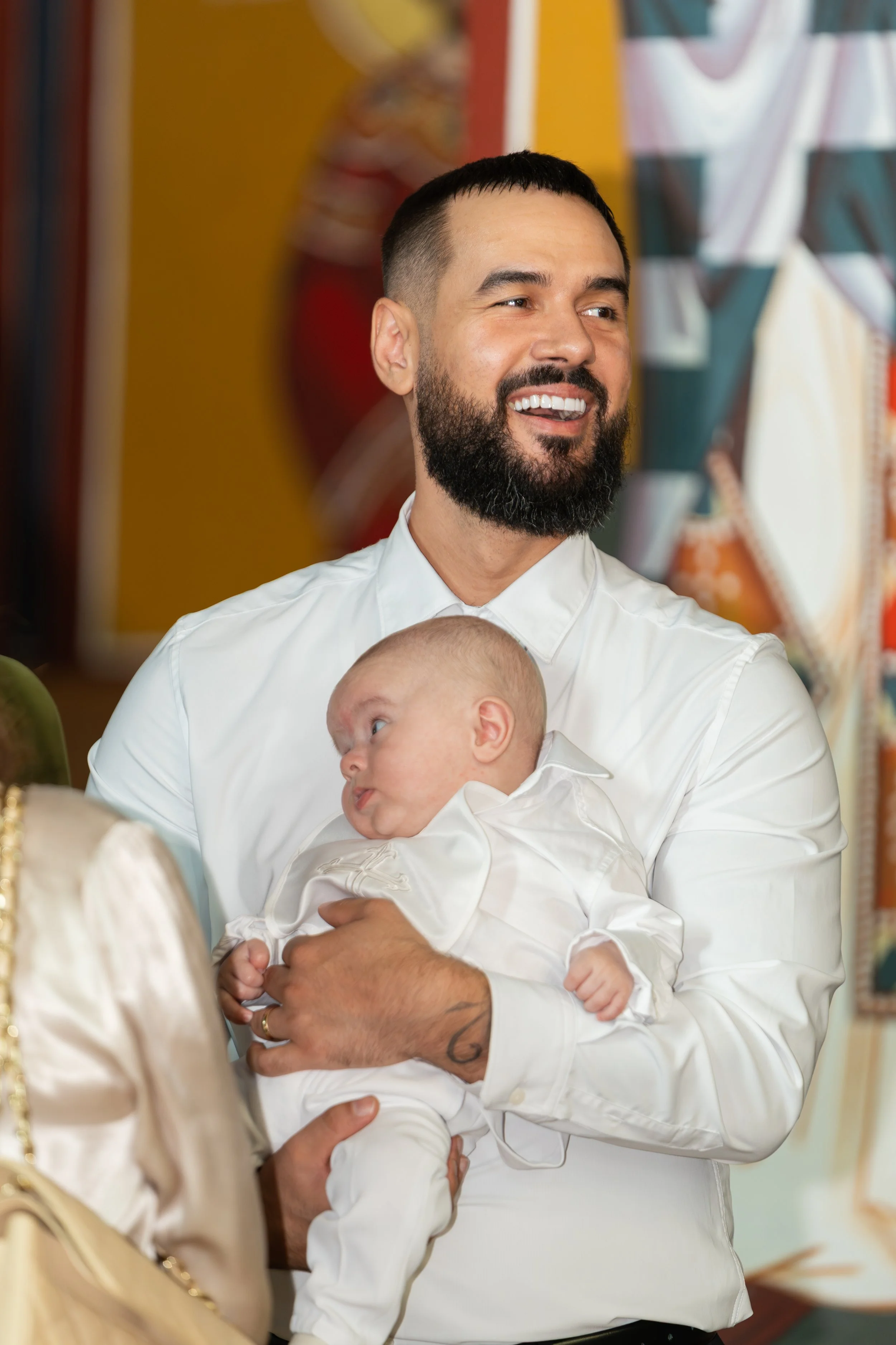 A man with a beard and short black hair smiling while holding a baby dressed in white. They are indoors with colorful artwork in the background.