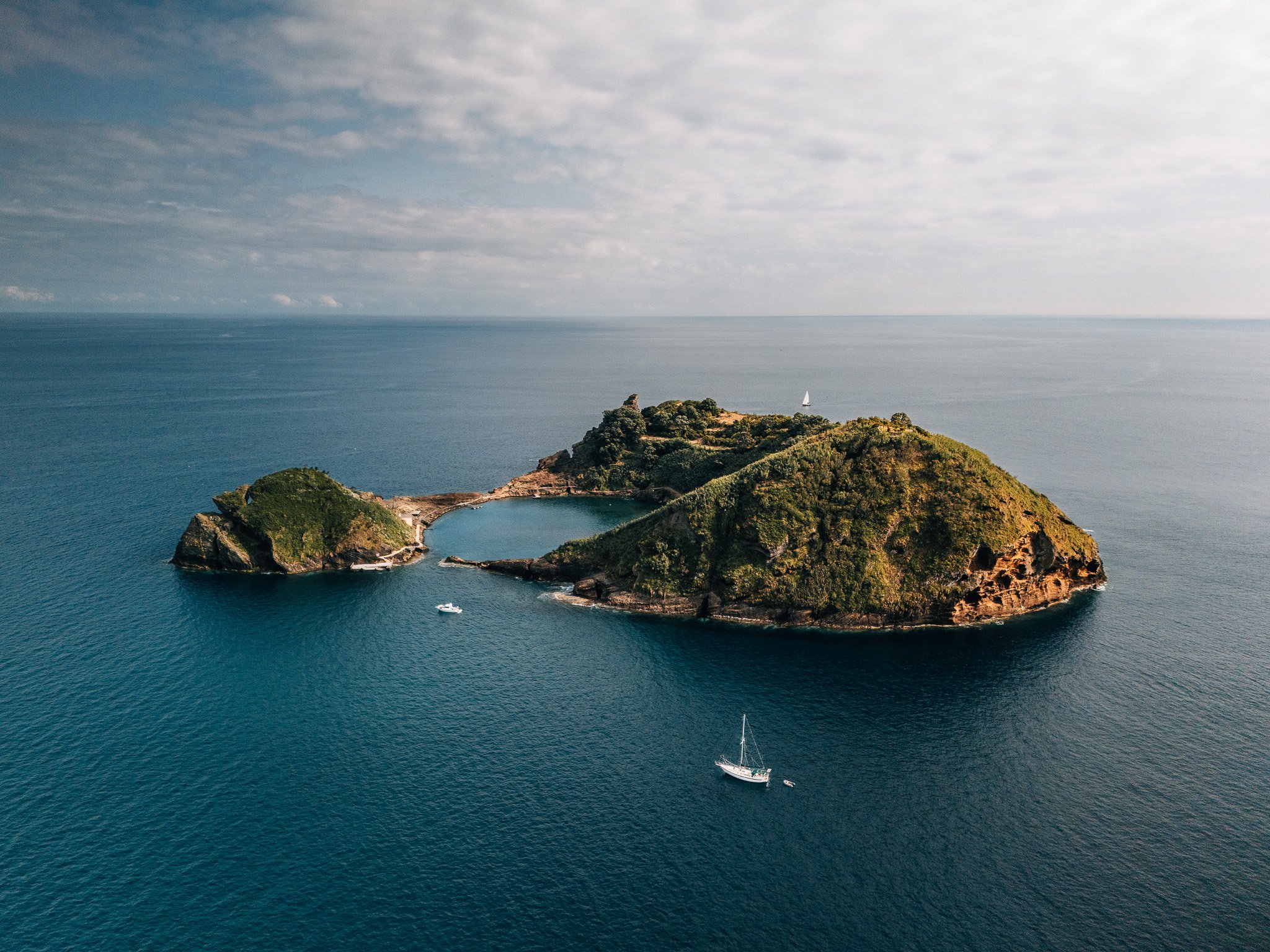 An aerial view of a small island with lush green trees and a rocky shoreline, surrounded by blue ocean waters with a sailboat nearby.