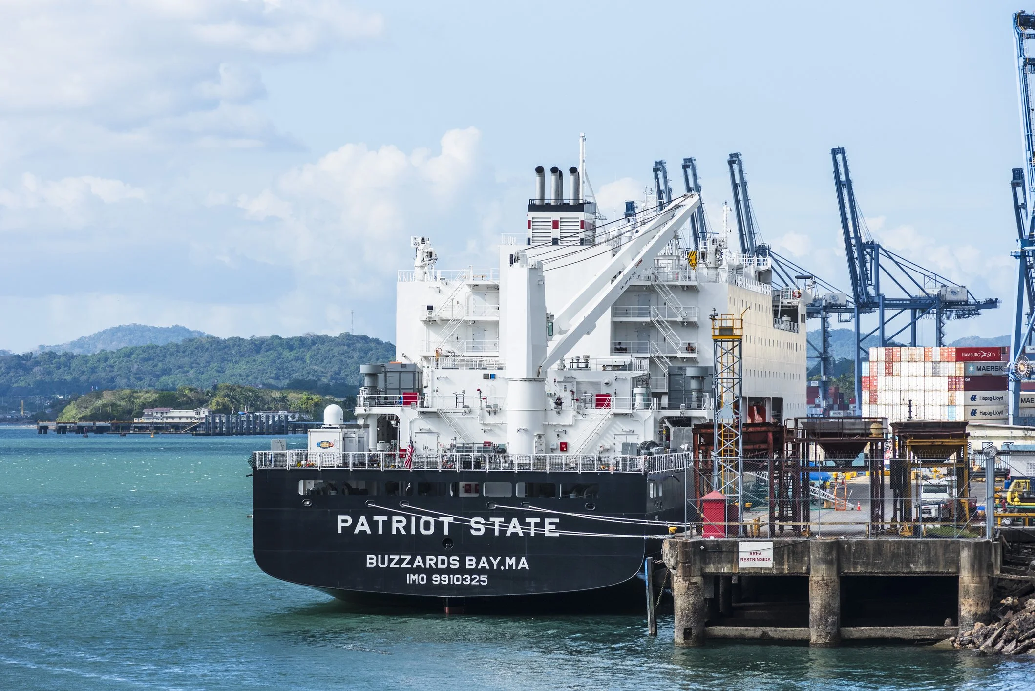 A large cargo ship named 'Patriot State' docked at a port with multiple cranes and containers in the background, with a hilly landscape and partly cloudy sky.