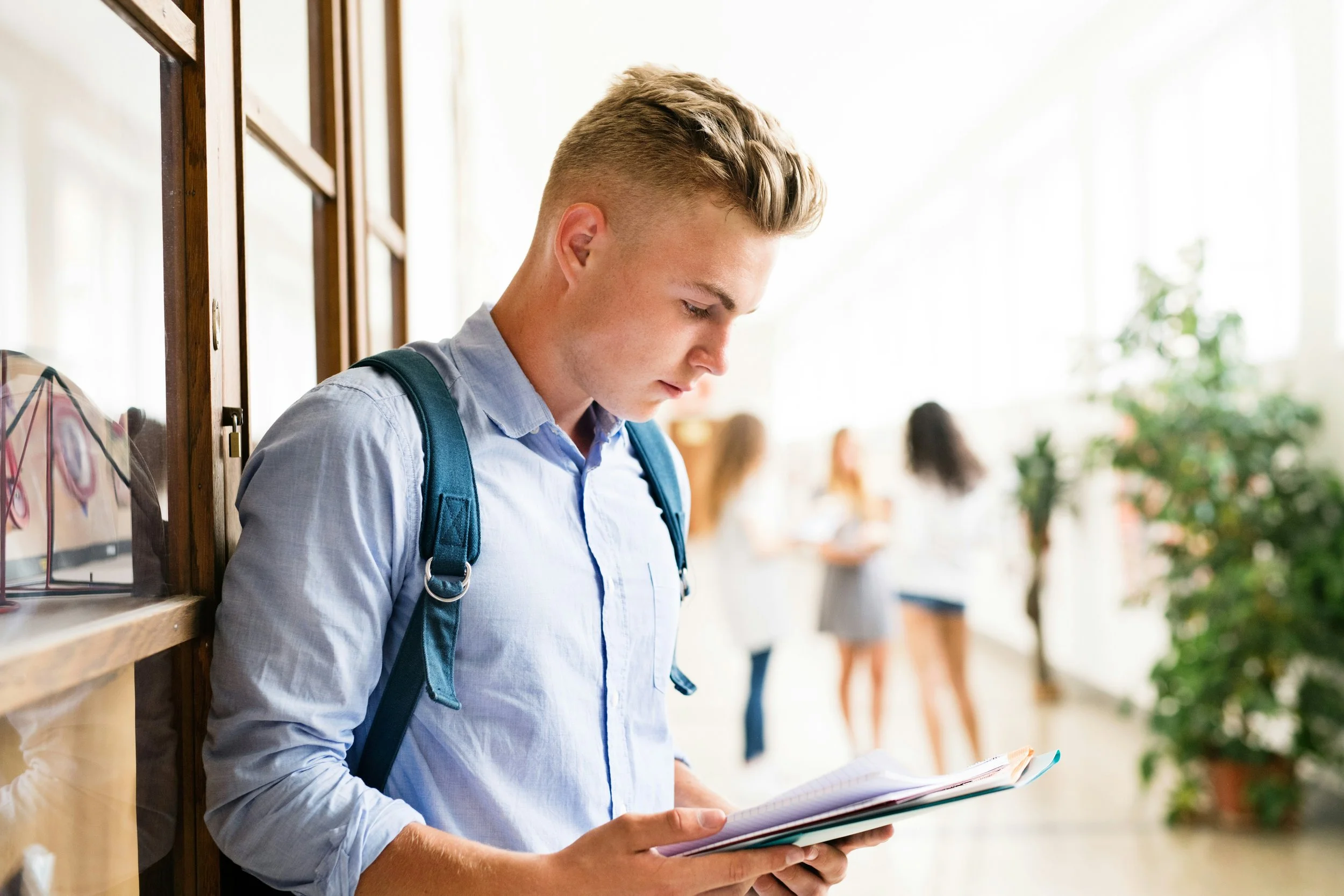 A young man with blond hair, wearing a light blue shirt and a backpack, leaning against a wooden wall while reading a notebook in a bright school hallway with other students in the background.