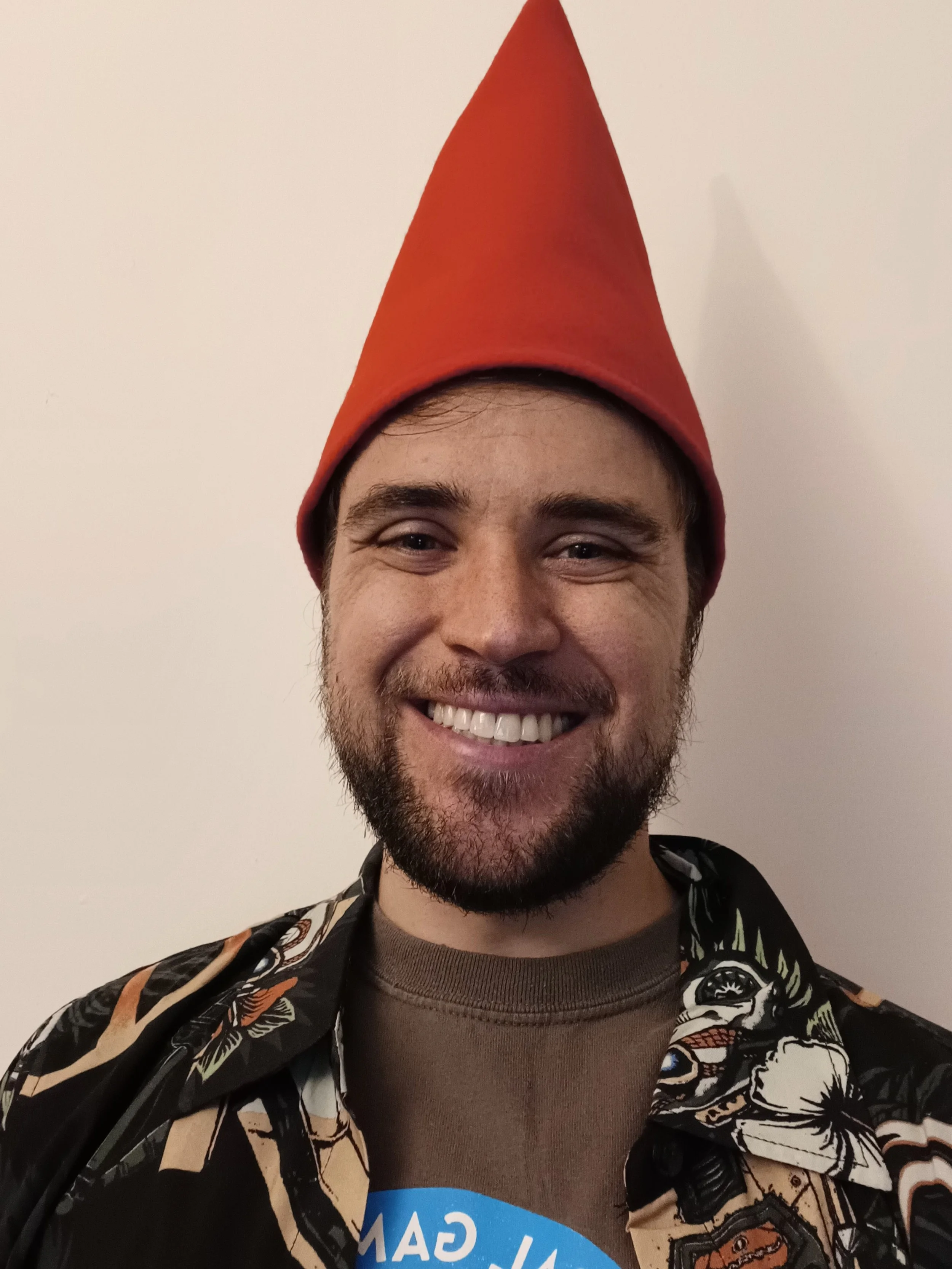 A smiling man with a beard wearing a red cone-shaped hat, a dark shirt with a colorful print, and a gray T-shirt underneath.