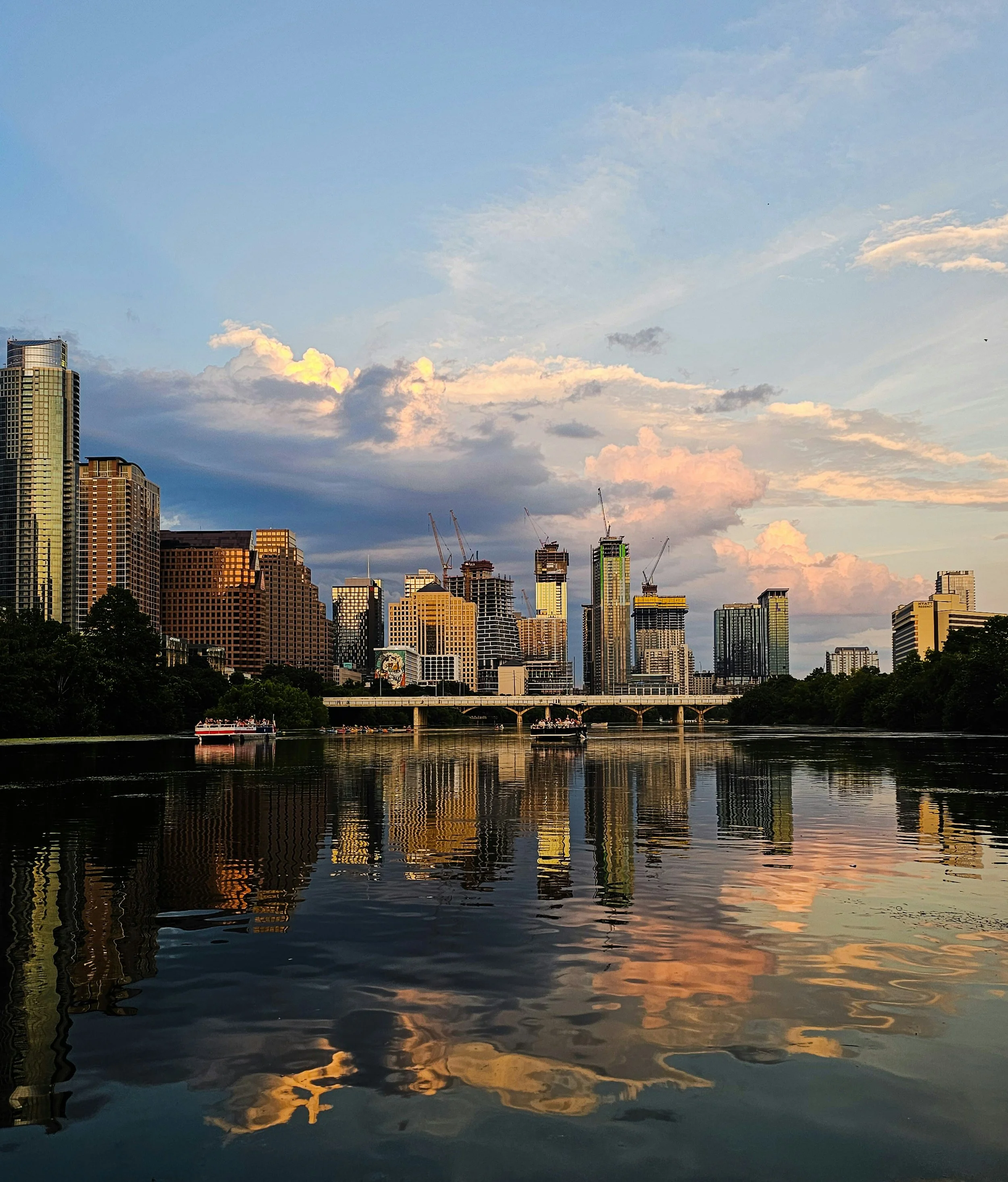 City skyline with tall buildings next to a body of water reflecting the buildings and partly cloudy sky at sunset.