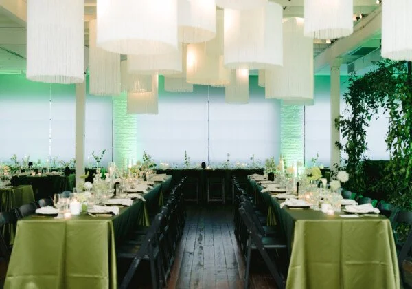 Elegant banquet room with long tables covered in green tablecloths, set with white plates and glassware, decorated with candles and small floral arrangements, illuminated by large hanging white lamps and surrounded by green plants.
