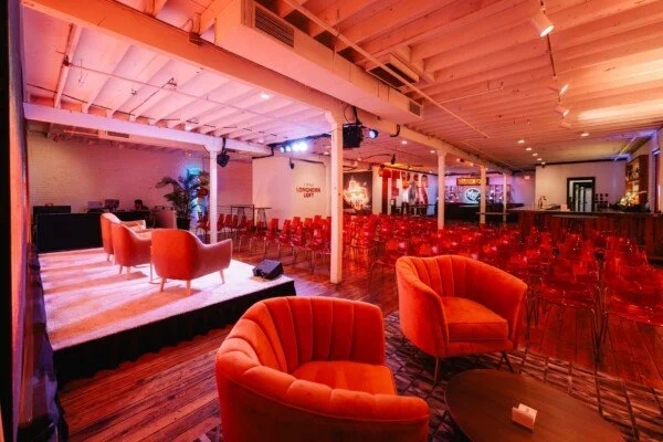 A cozy event space with pink and purple lighting, featuring a small stage with two pink chairs, a large room with red chairs, and sofas near the stage, decorated with plants and vintage posters.