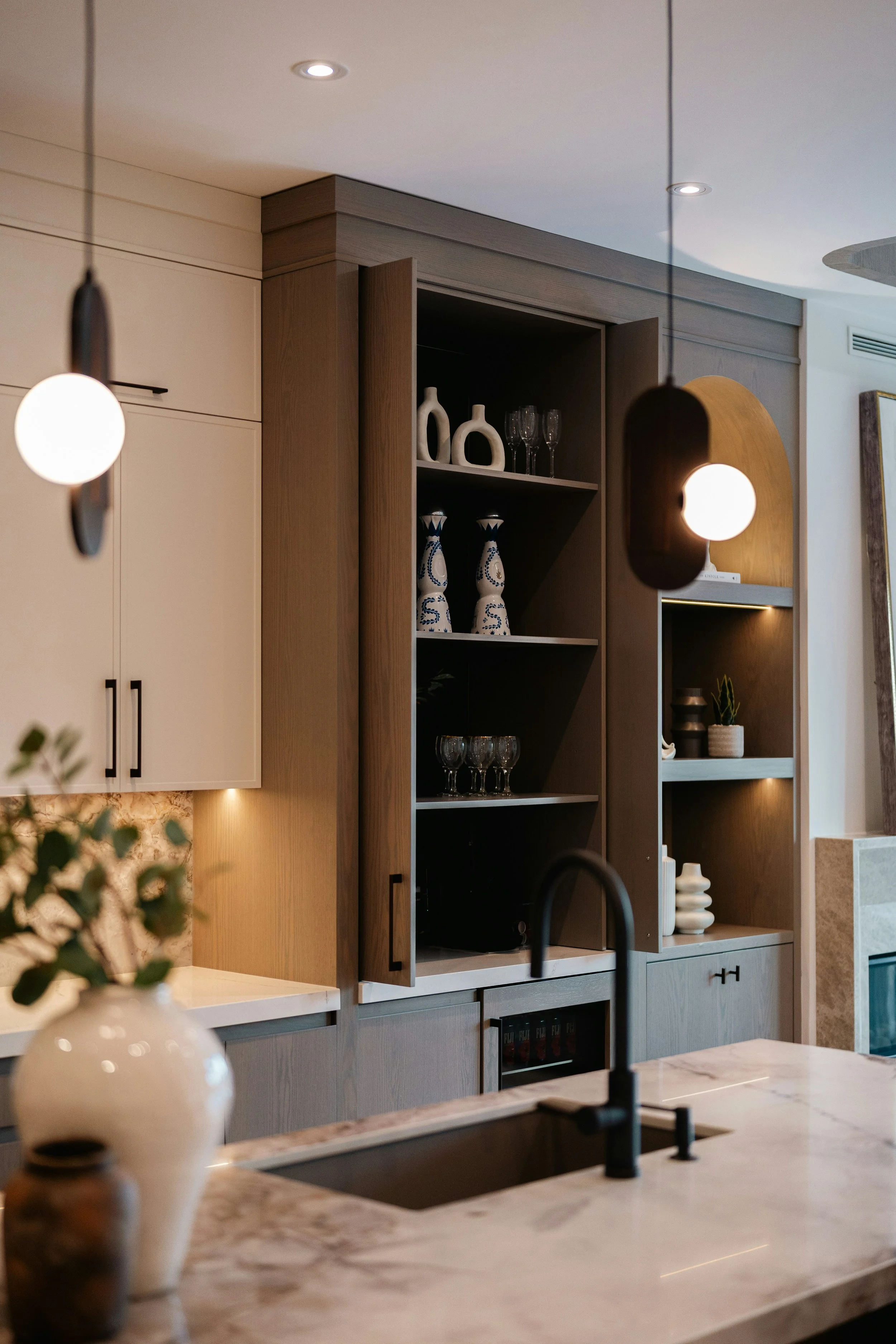 Modern kitchen with beige and wood cabinetry, open shelves displaying glassware and decorative vases, marble countertop, black faucet, and pendant lighting.