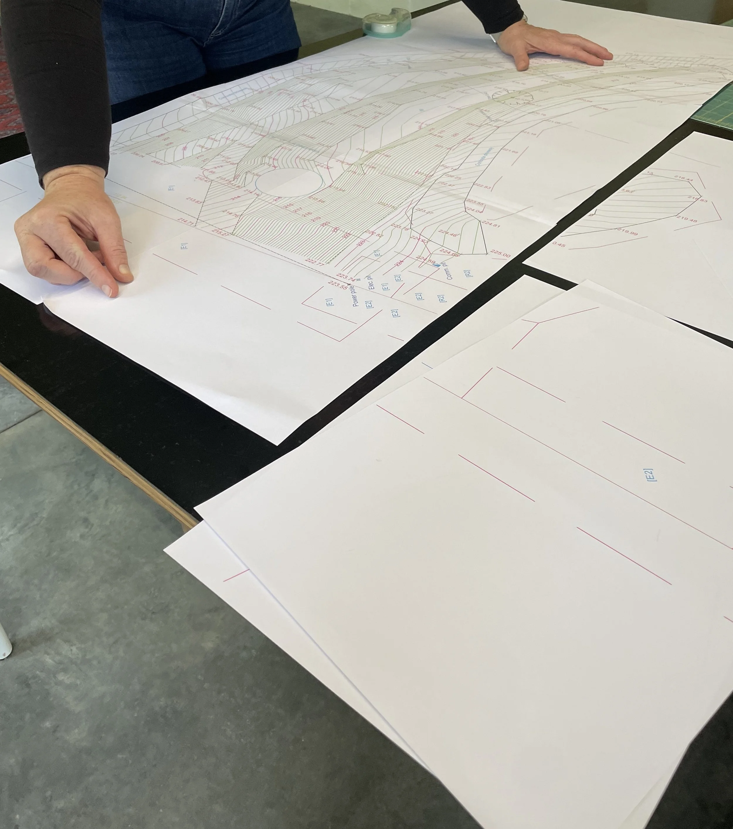 A person is examining an enlarged technical or architectural drawing on a large white sheet of paper laid out on a table. The person highlights details, and the diagram includes colored lines, measurements, and annotations.
