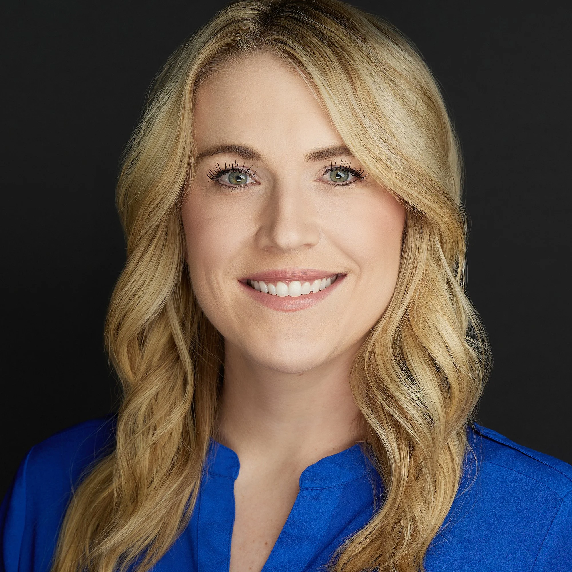 Portrait of a smiling woman with blonde wavy hair, wearing a blue blouse, against a dark background.